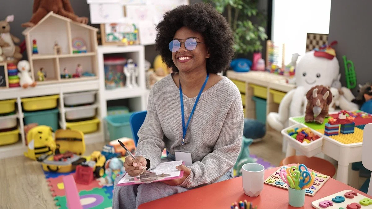 A smiling woman with glasses and curly hair sitting at a table in a colorful classroom with toys and learning materials.