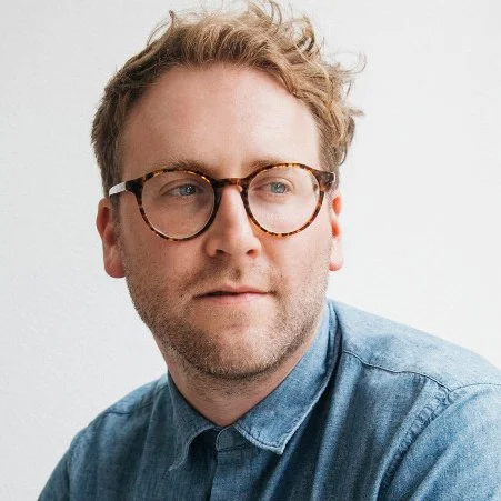 A young man with light brown hair and glasses, wearing a denim shirt, looking to the side against a light background.