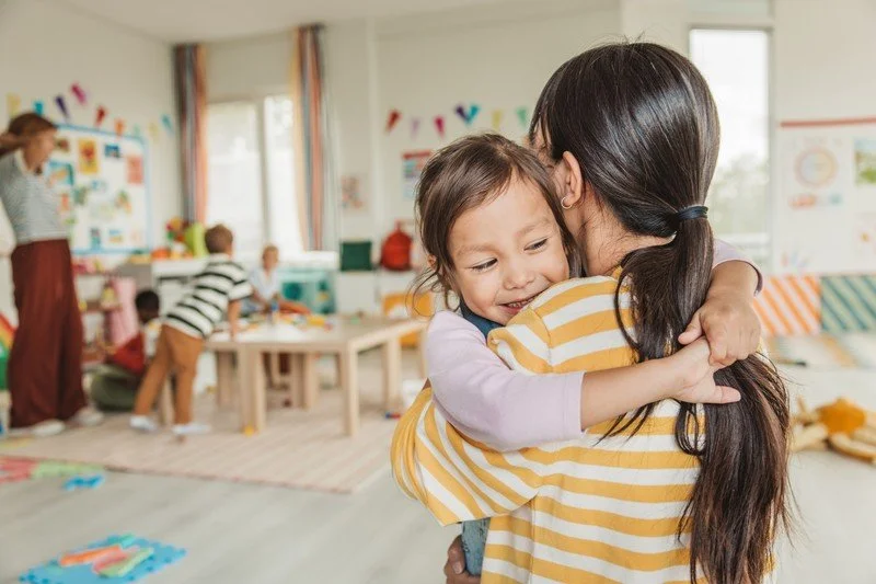 A woman hugging a smiling child in a colorful classroom with other children and an adult in the background.