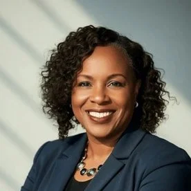 Professional woman with curly hair smiling, wearing a navy blazer and necklace.