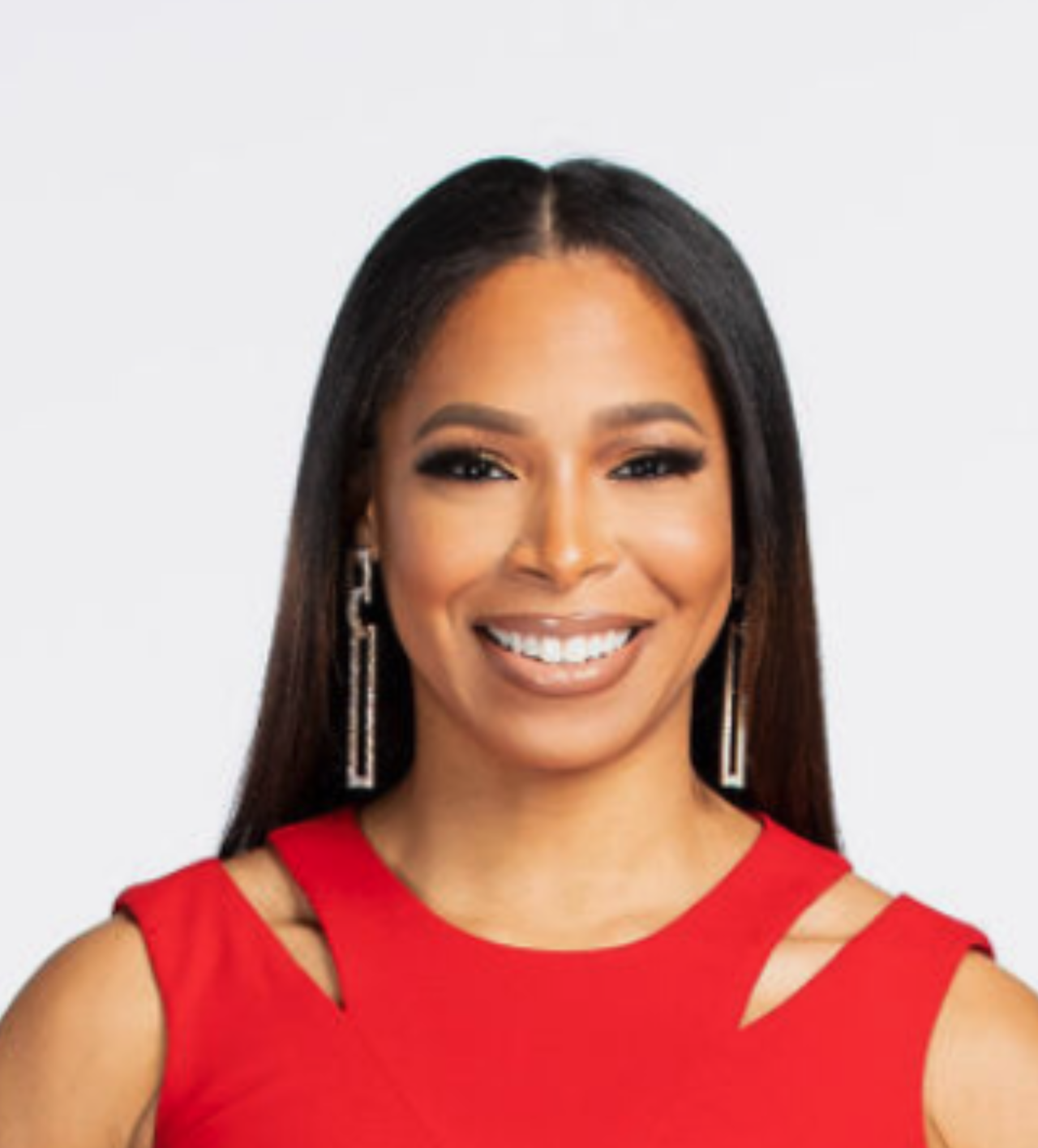 Smiling woman with short dark hair wearing a blue sweater with white flower details and a delicate necklace, standing against a plain white background.