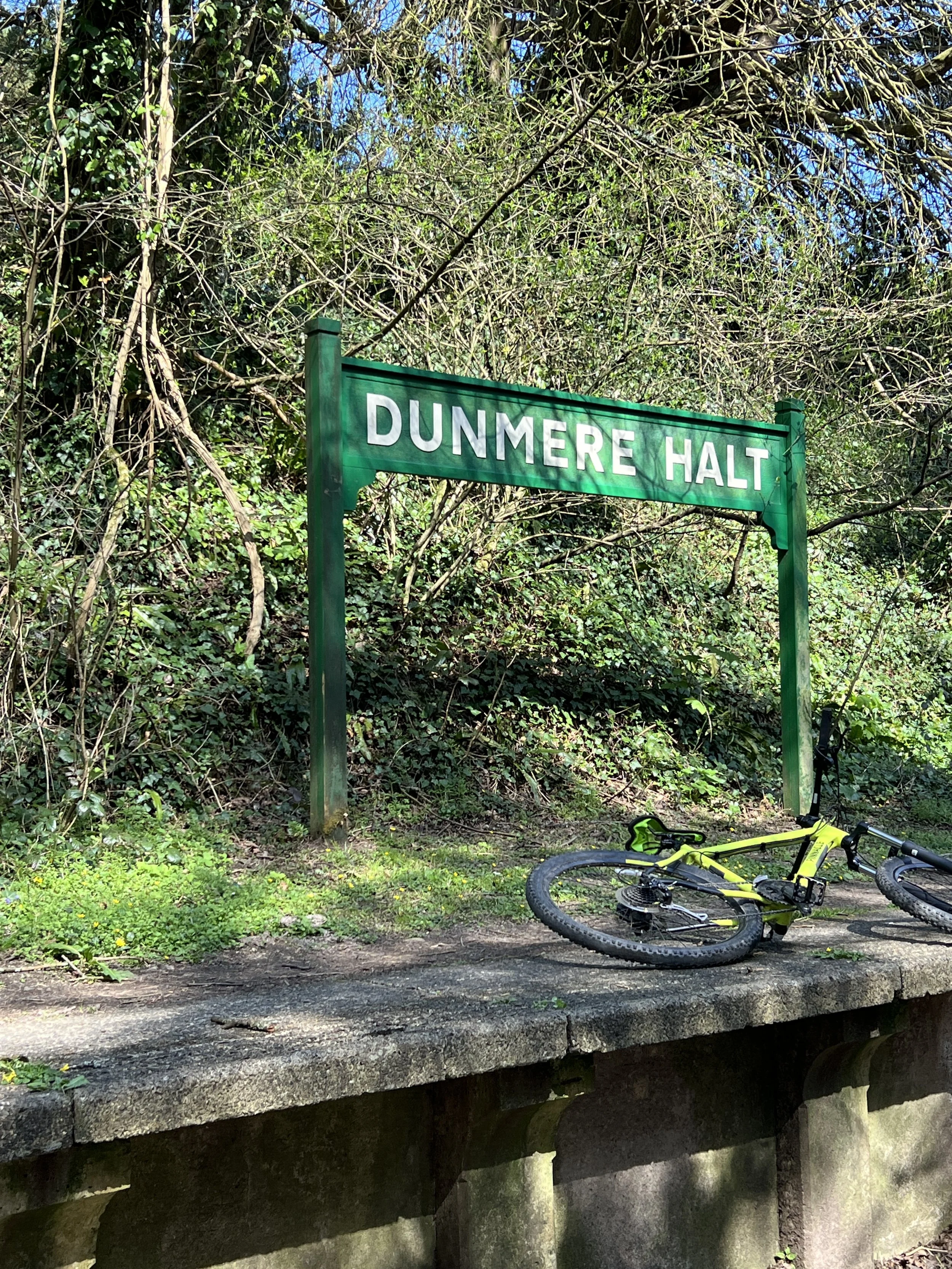 A green street sign that reads 'DUNMERE HALT' is mounted on two posts next to a bicycle lying on the ground. The area is surrounded by dense greenery and trees.