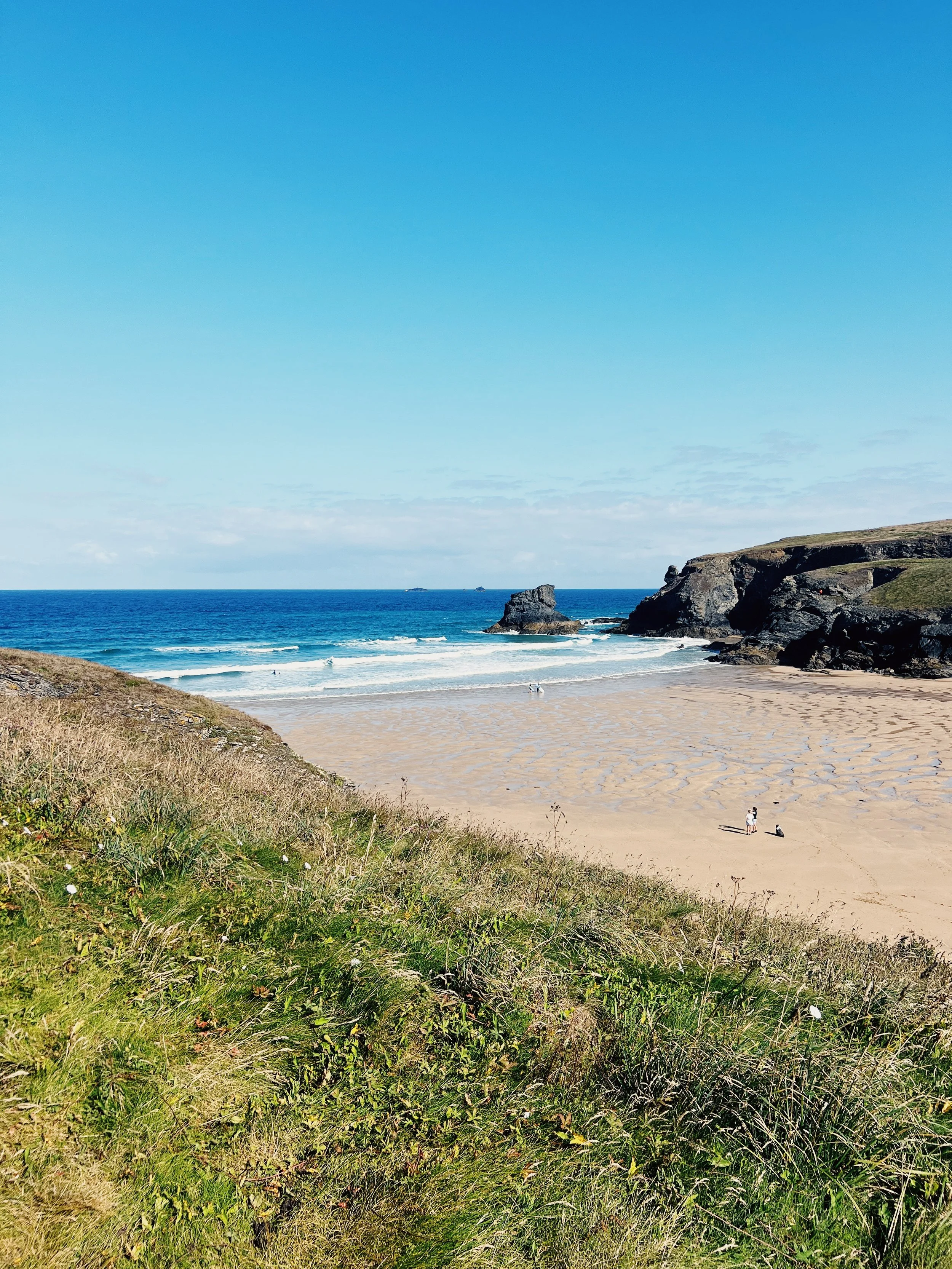 Sandy beach with grassy dunes in the foreground, rock formations in the distance, and the ocean with gentle waves under a bright blue sky.