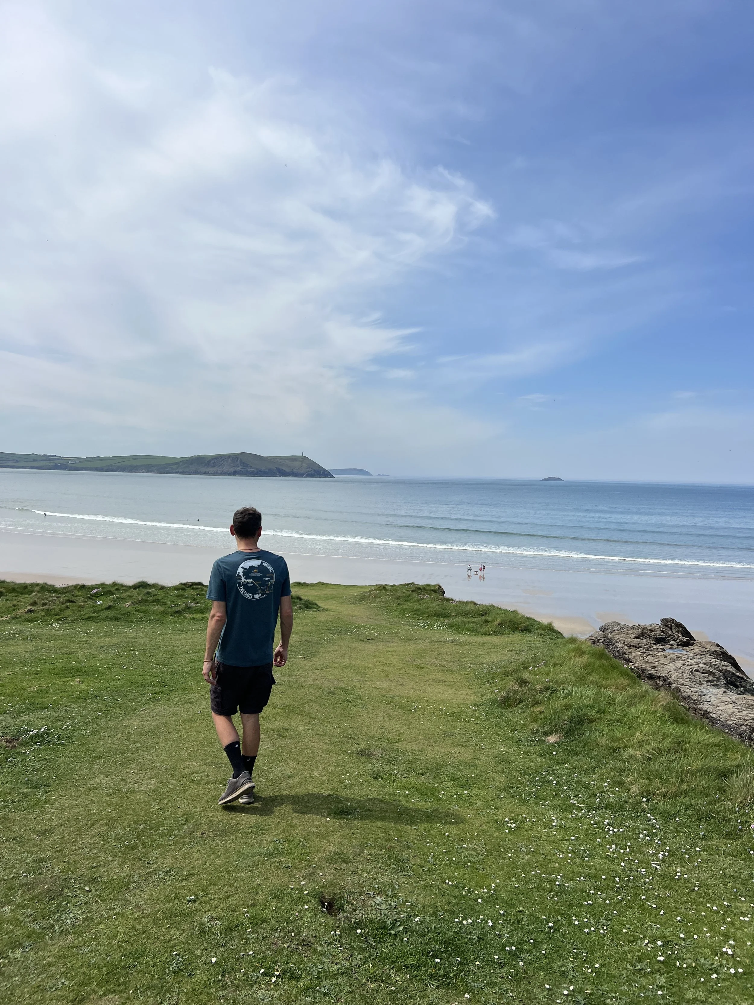 A person walking on a grassy path near the beach with the ocean and a partly cloudy sky in the background.