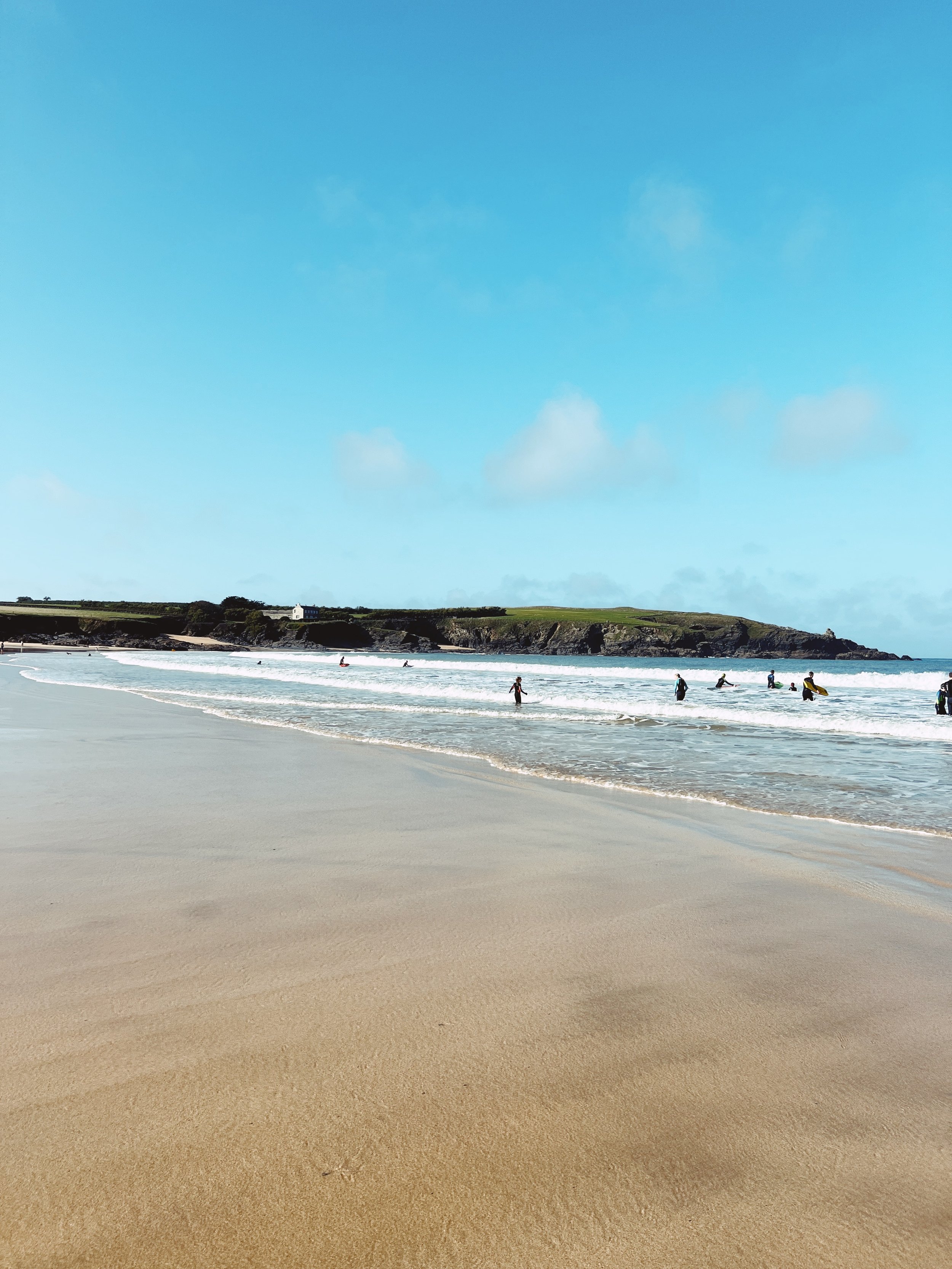 People swimming and surfing at a beach with golden sand, ocean waves, and a hillside in the background under a partly cloudy blue sky.