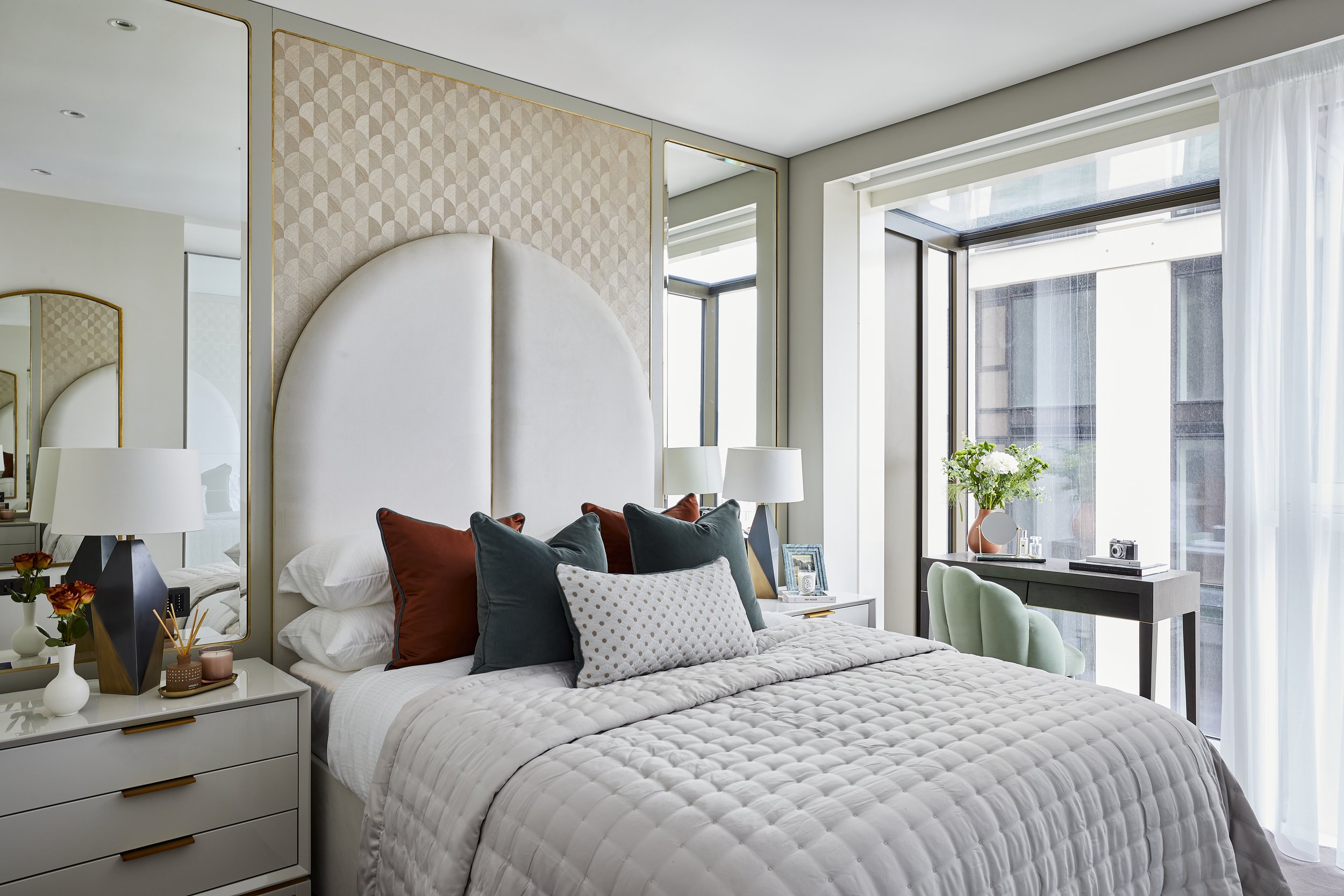 A modern bedroom with a large bed covered in a quilt, multiple pillows, and a white with decor including vases, a lamp, and personal items. There are large windows with sheer white curtains letting in natural light, a desk with a chair, flowers, and a small mirror, and a headboard with textured wallpaper.