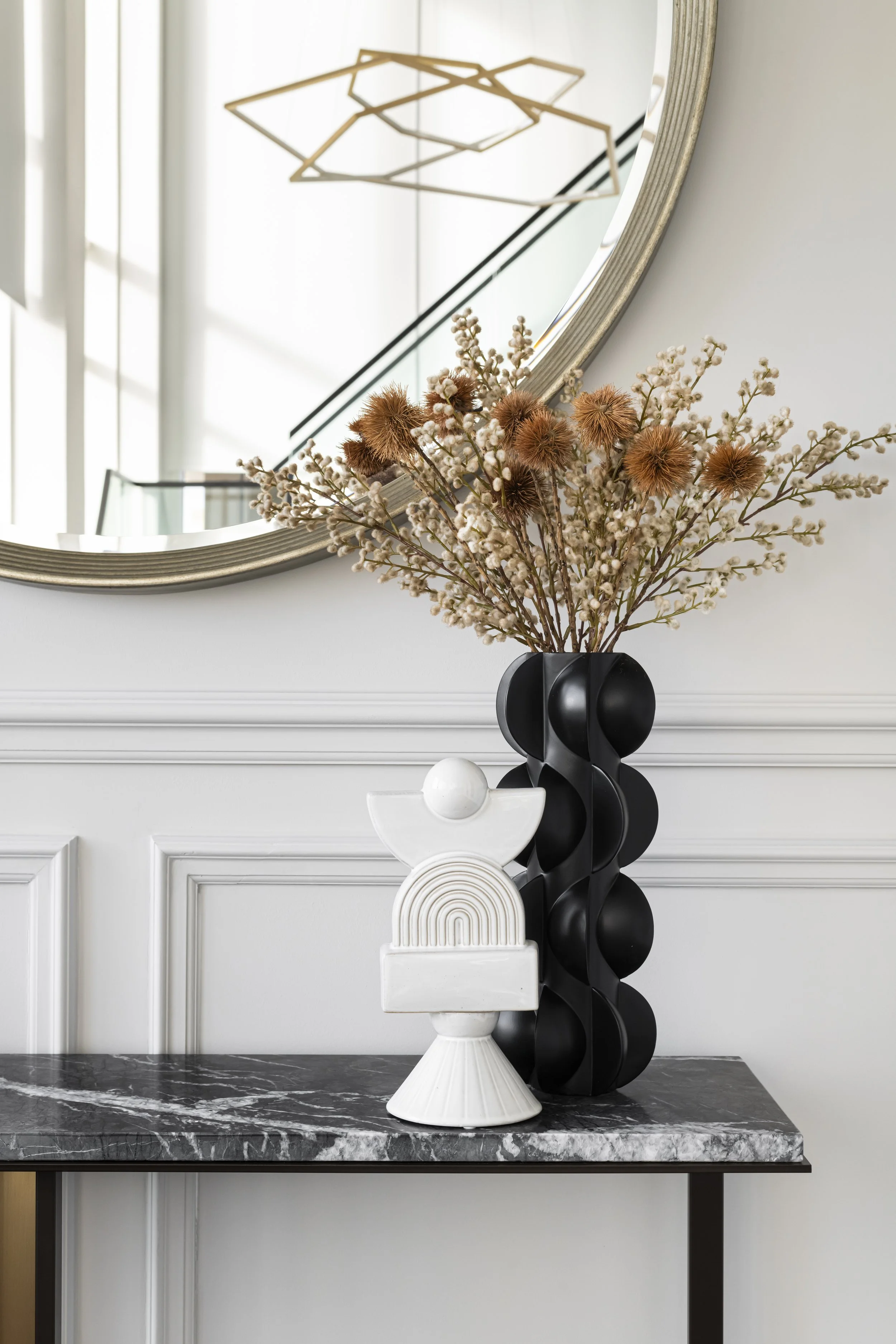 A modern interior decor scene featuring a round mirror with a silver frame, a geometric chandelier reflected in the mirror, a black vase with dried flowers, a white sculptural vase, and a marble tabletop against a white wall with decorative molding.