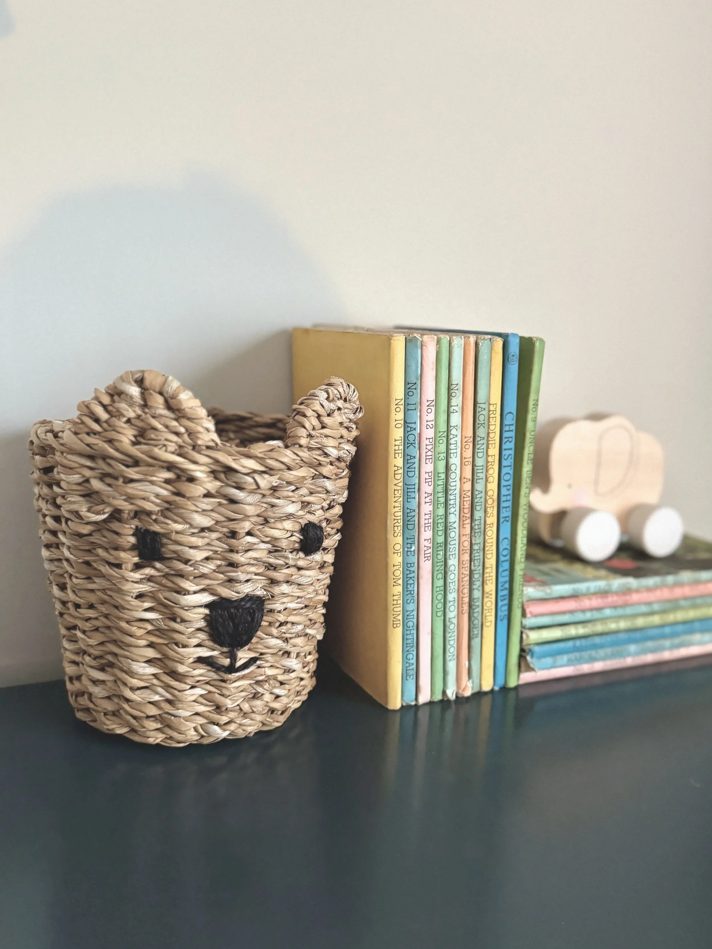 A woven basket with a bear face design next to a stack of colorful children's books on a black surface.