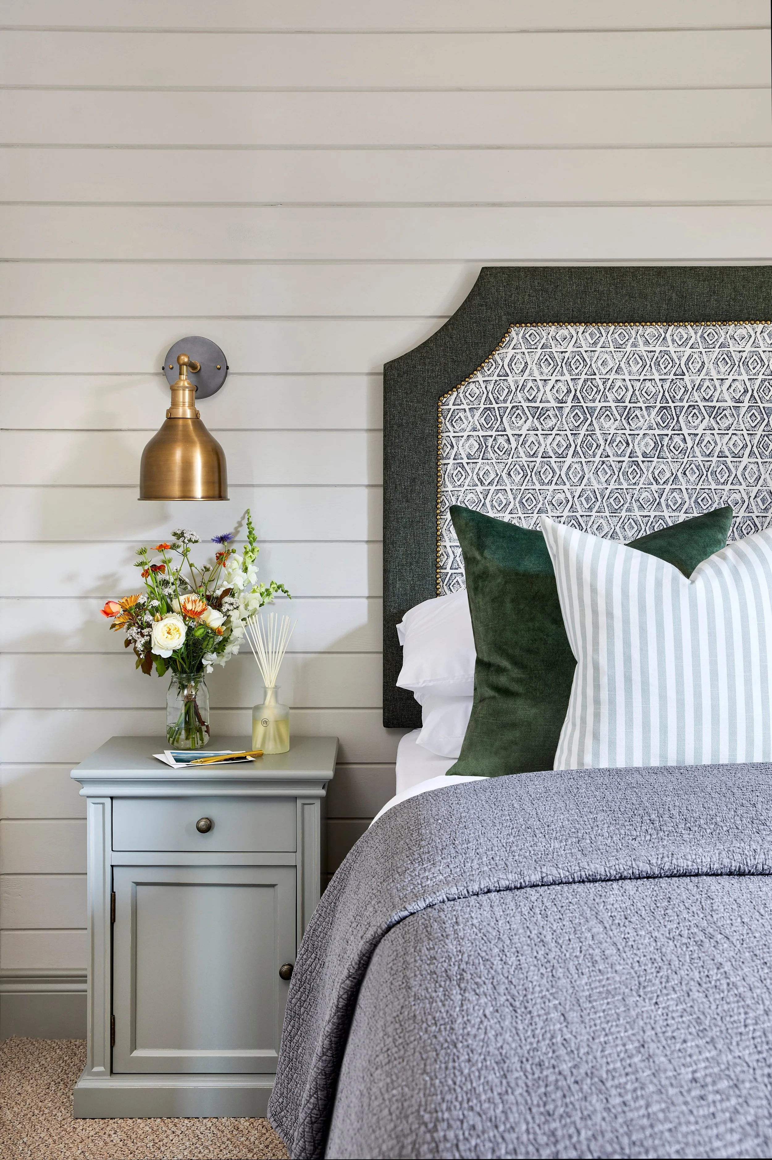 A bedroom scene featuring a bedside table with a drawer, decorated with a bouquet of flowers and a bottle of perfume. To the right, part of a bed with white sheets, three pillows with green and white striped covers, a gray quilted coverlet, and a hea