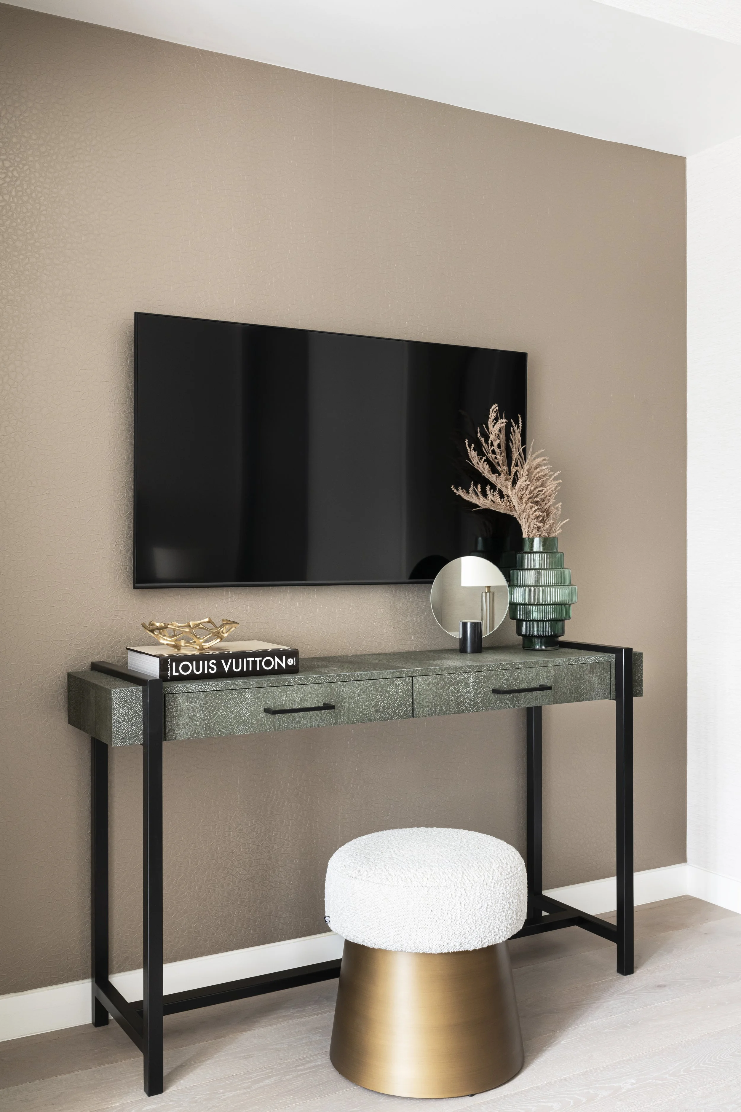 Living room with a wall-mounted flat-screen TV above a dark green console table decorated with a gold decorative object, a book titled Louis Vuitton, and a green vase holding pink pampas grass. In front is a white textured rounded stool with a gold b