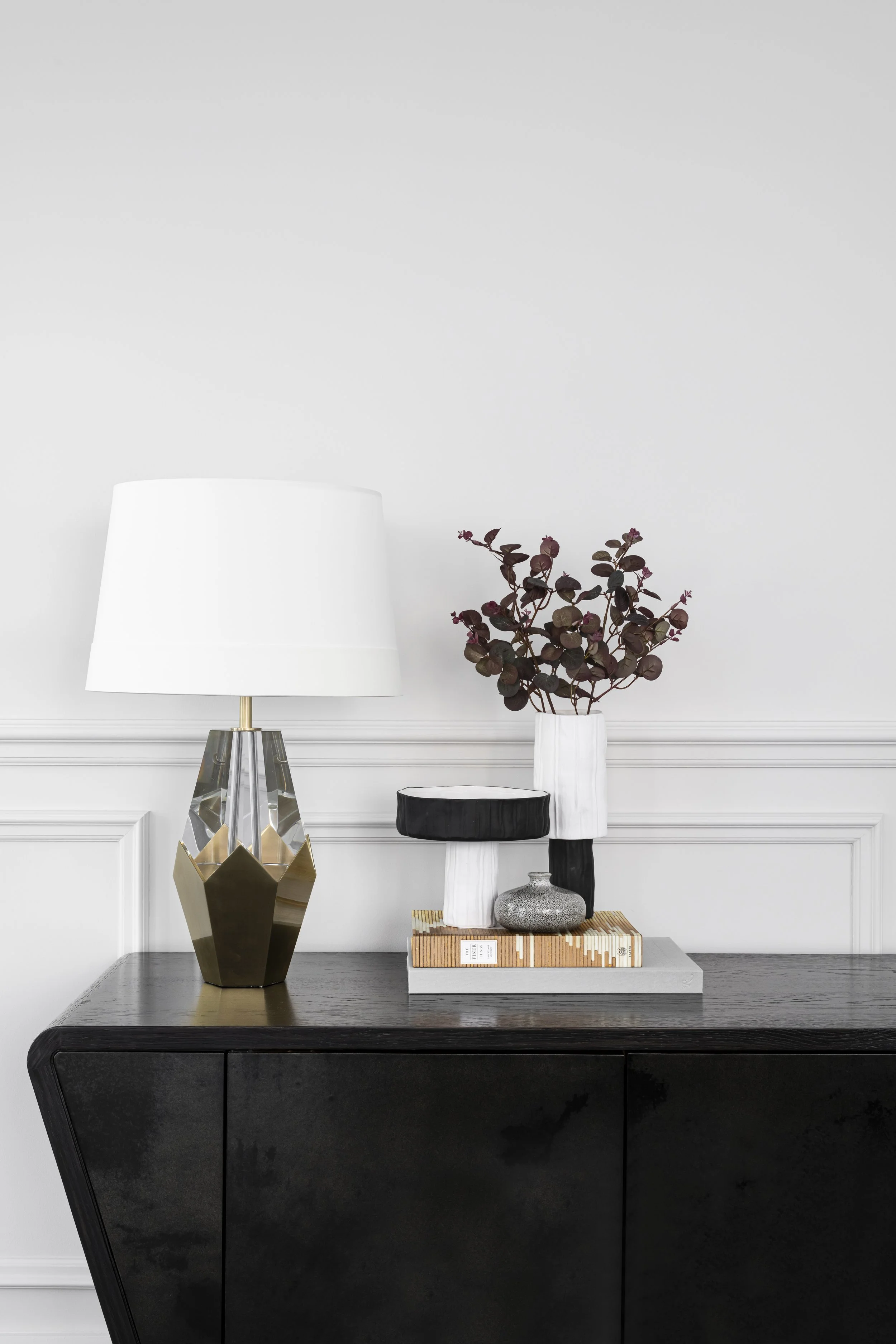 A modern black sideboard, decorated with a gold and glass geometric lamp, a white vase with dark red and green leaves, and a small grey stone sculpture, on a wooden tray. In the background, a white wall with decorative moulding.