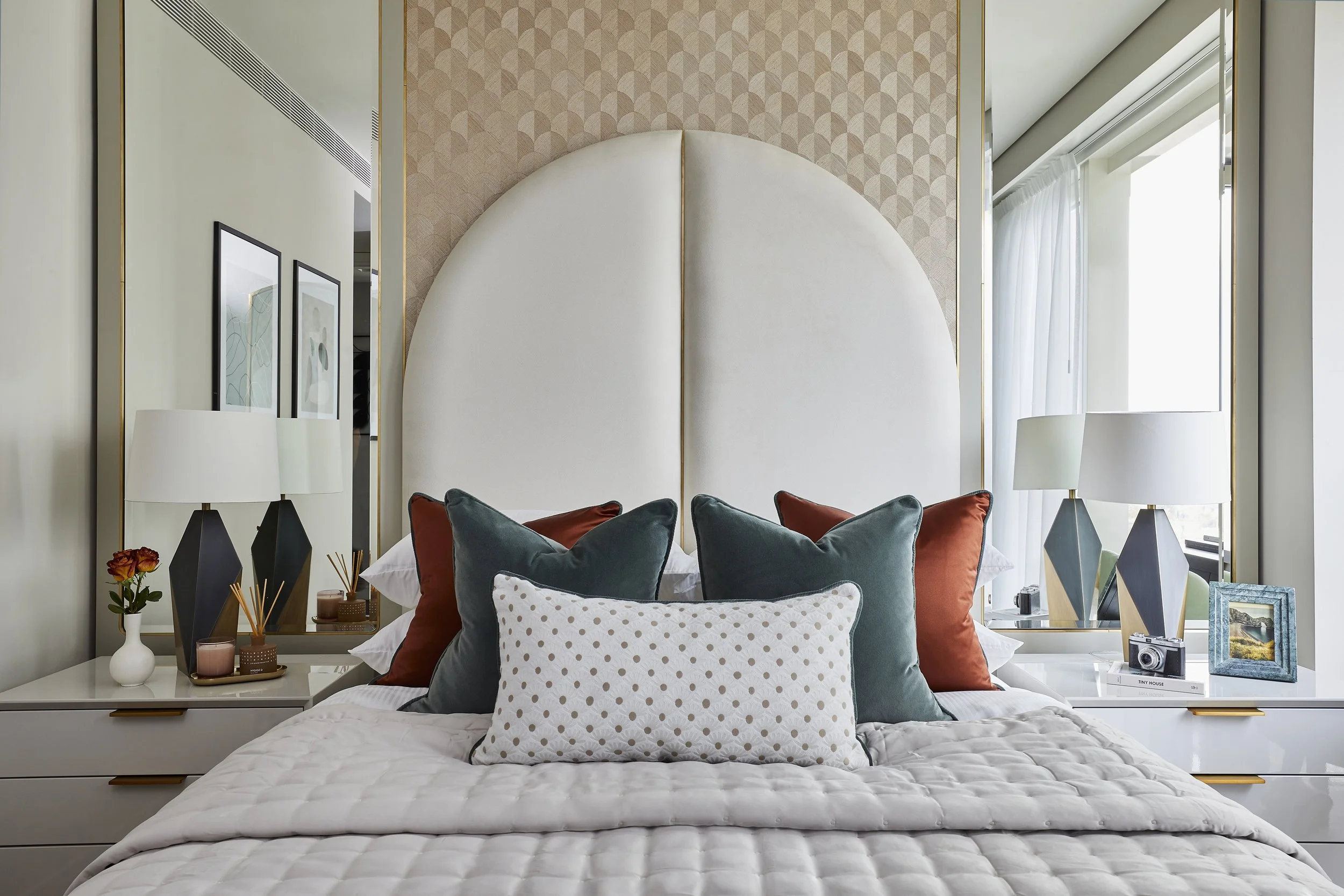 Modern bedroom with white quilted bed, array of decorative pillows, white bedsides, table lamps, framed artwork, large window with sheer curtains, and a decorative textured geometric headboard.