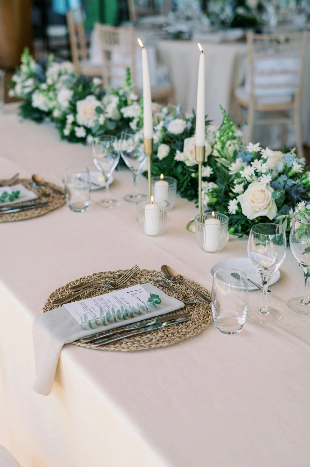 Elegant wedding or event table setting with a beige tablecloth, woven placemats, silverware, glassware, a menu, and a floral centerpiece with white and green flowers, candles, and gold candle holders.