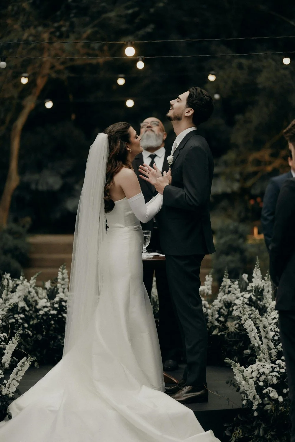 Wedding ceremony outdoors with bride, groom, and officiant under string lights, surrounded by white flowers.