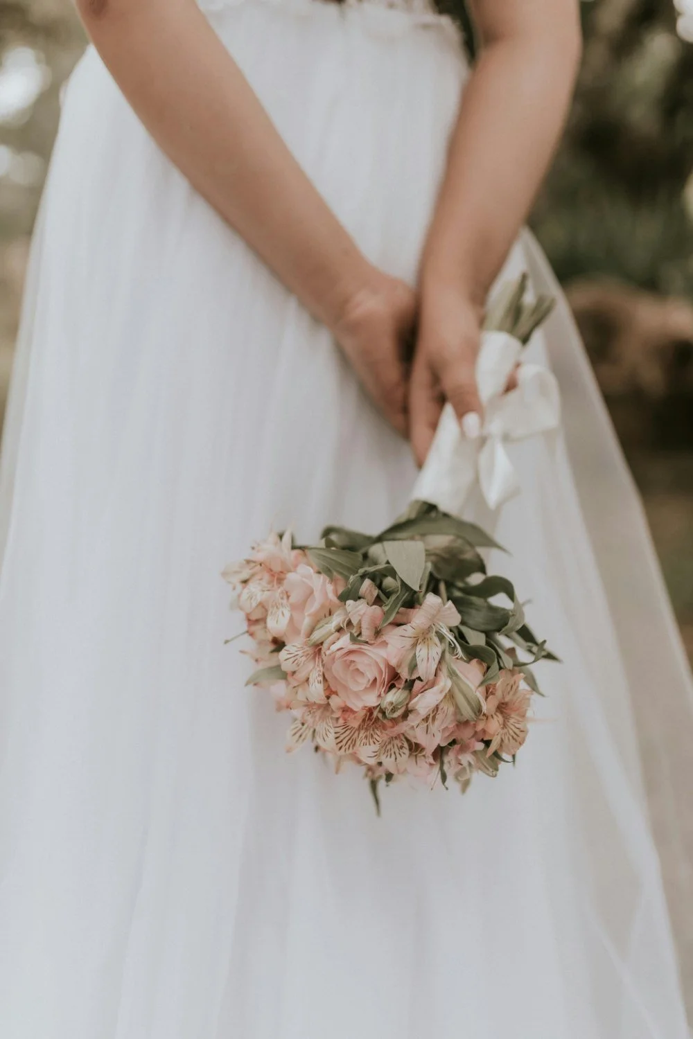 A bride in a white dress holding a bouquet of pink roses, lilies, and greenery behind her back.