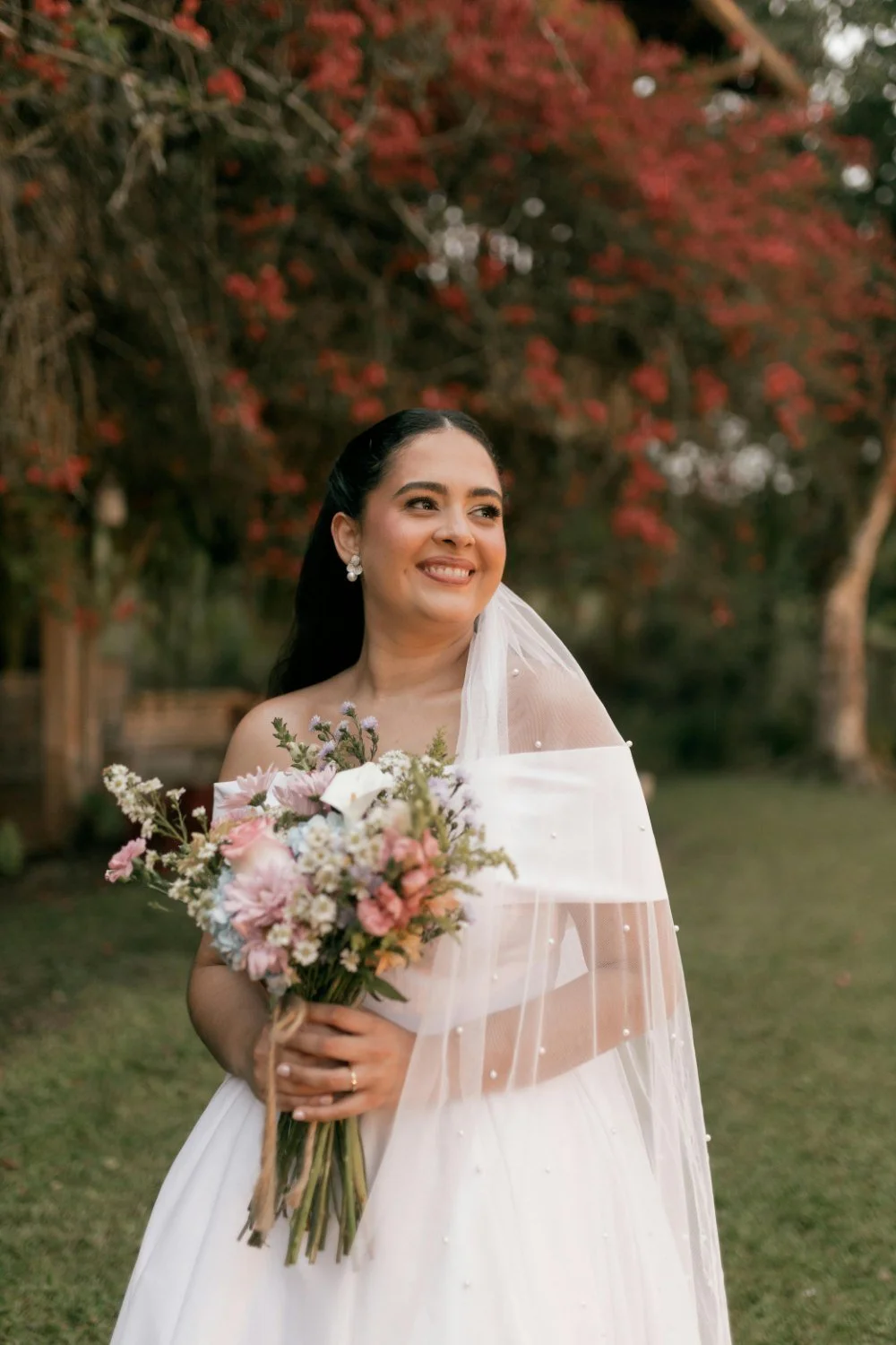 A bride standing outdoors, smiling, holding a bouquet of pink and white flowers, with trees and red foliage in the background.