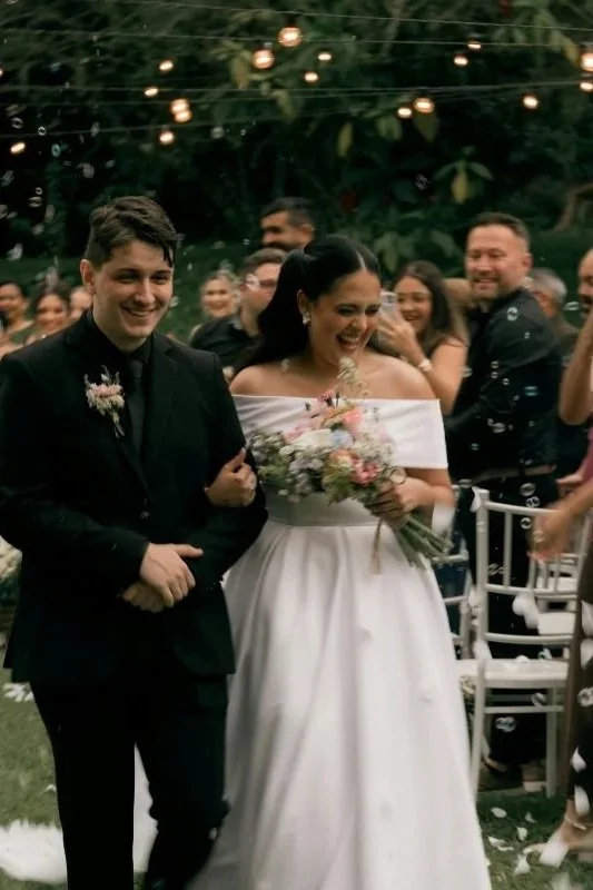 A bride and groom walking arm in arm at their outdoor wedding reception, surrounded by smiling guests and hanging string lights.