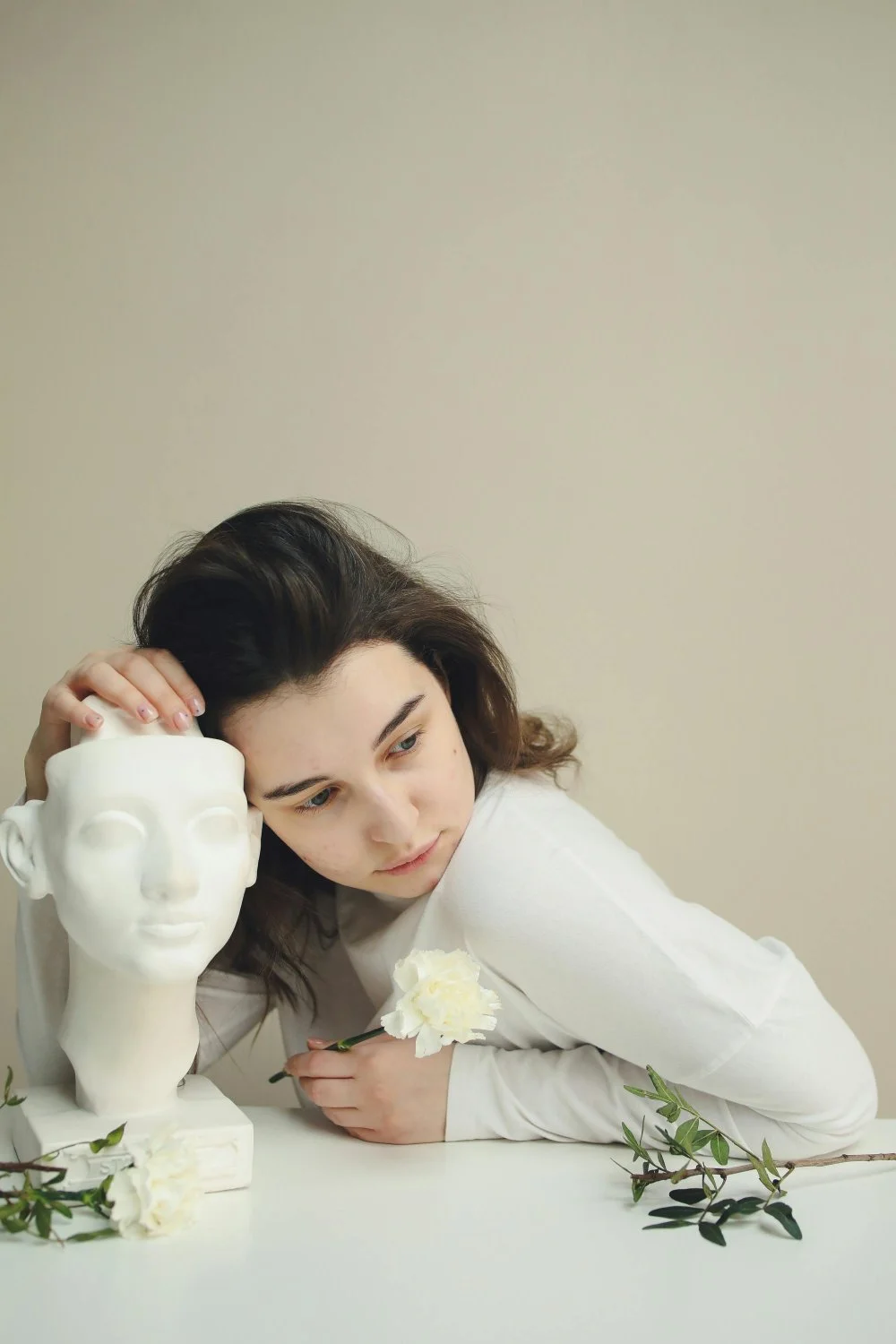 Young woman with dark hair leaning on a white table, resting her head on a white bust sculpture, holding a white flower, surrounded by green leaves, against a plain beige wall.