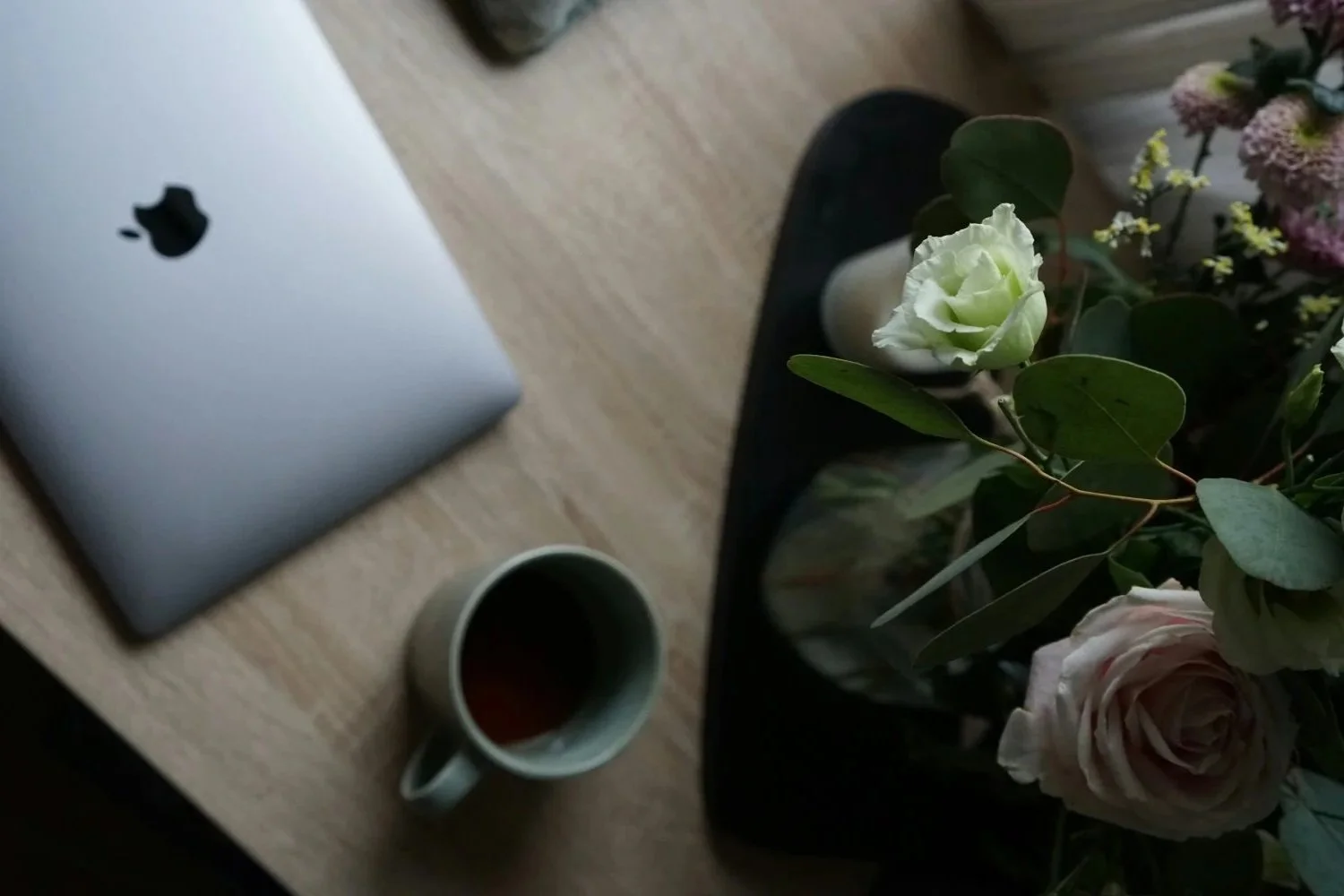 A wooden table with a silver MacBook, a cup of tea, and a bouquet of flowers in a vase.