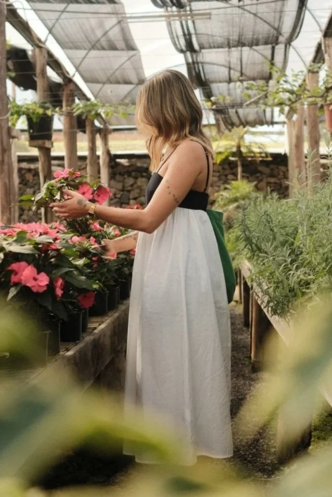 Woman with blonde hair wearing a sleeveless black and white dress, standing in a greenhouse inspecting pink flowers.