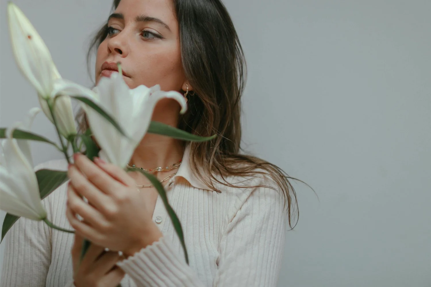 A woman with brown hair and light skin holding a bouquet of white lilies, wearing a cream ribbed cardigan and layered necklaces, looking to the side against a plain background.