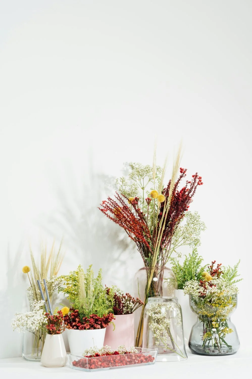Arrangement of various flowers in glass and ceramic vases on a white surface against a white background.
