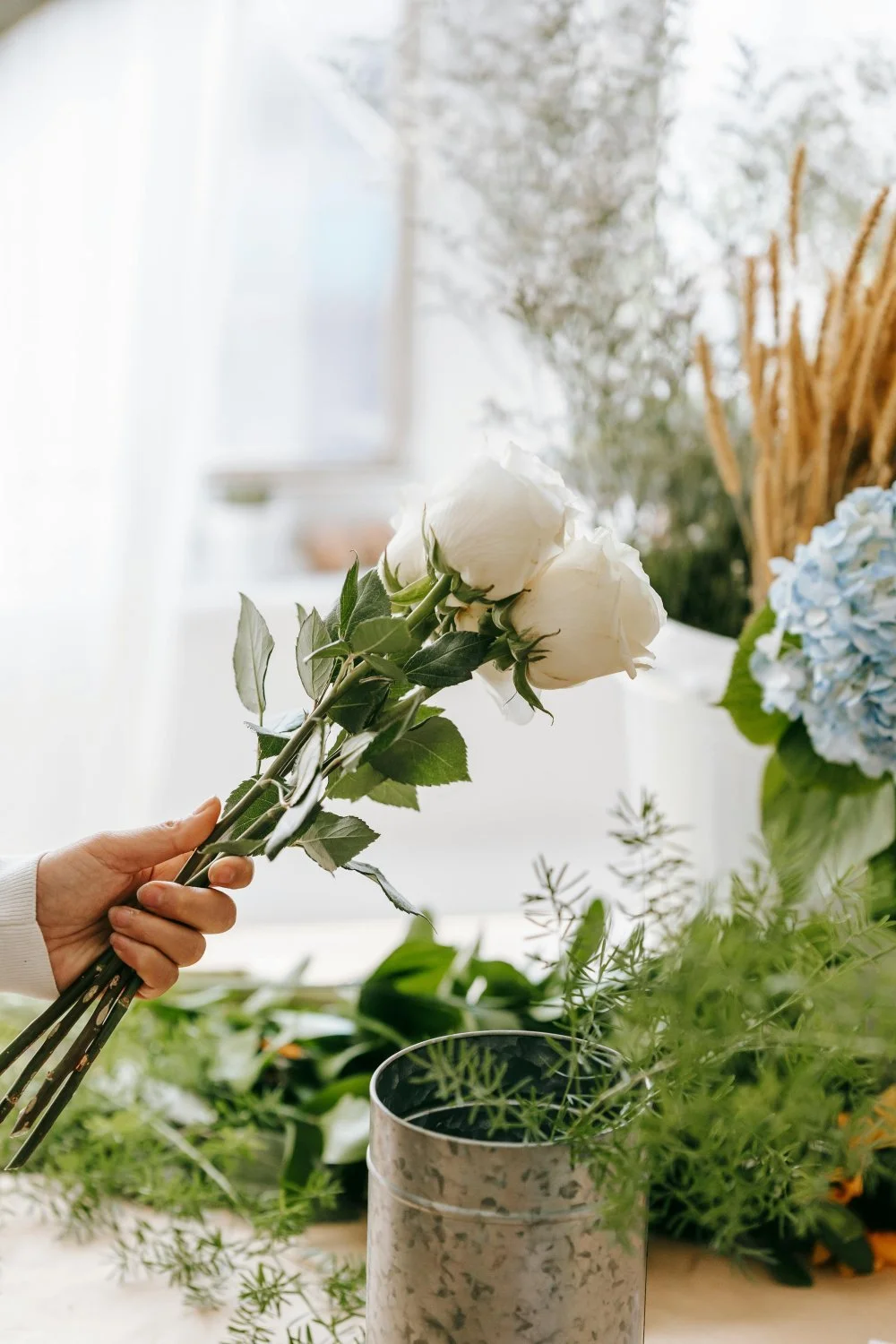 A person holding a bunch of white roses in a floral shop or flower arranging area filled with various flowers and greenery.
