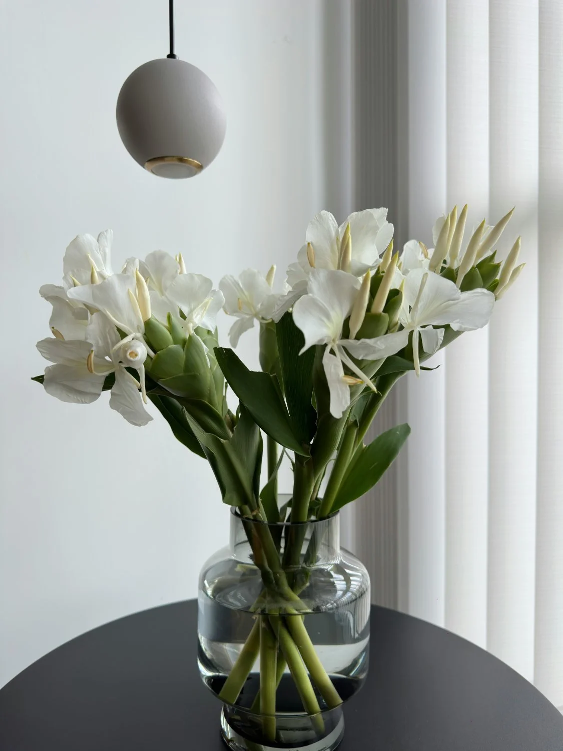 White flowers in a clear glass vase on a black table, with a modern spherical ceiling light and white curtains in the background.