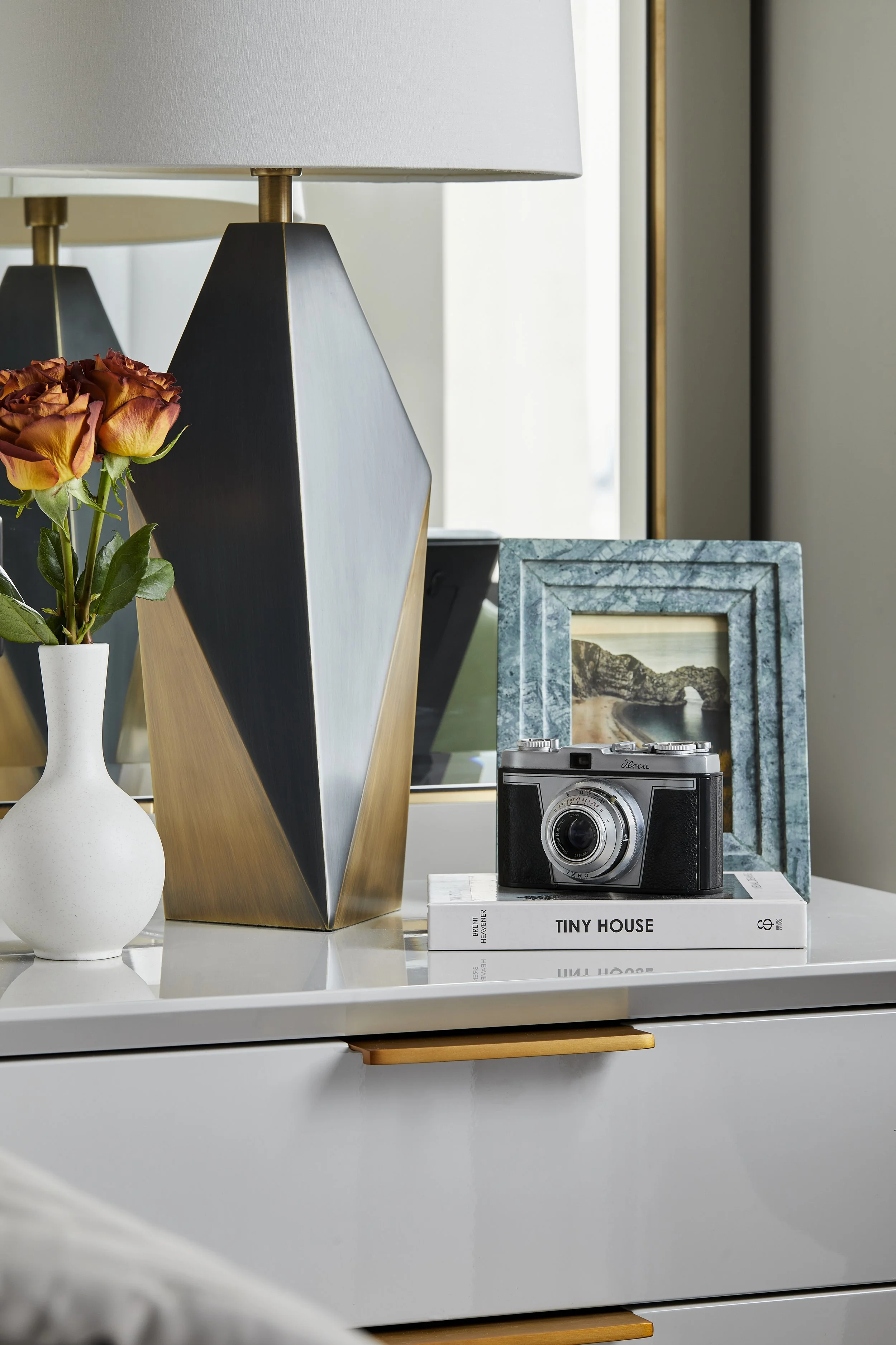 A white bedside table with a small white vase holding pink and orange roses, a geometric black and gold table lamp, a vintage camera, a framed landscape photo, and a book titled 'Tiny House' near a window with natural light.
