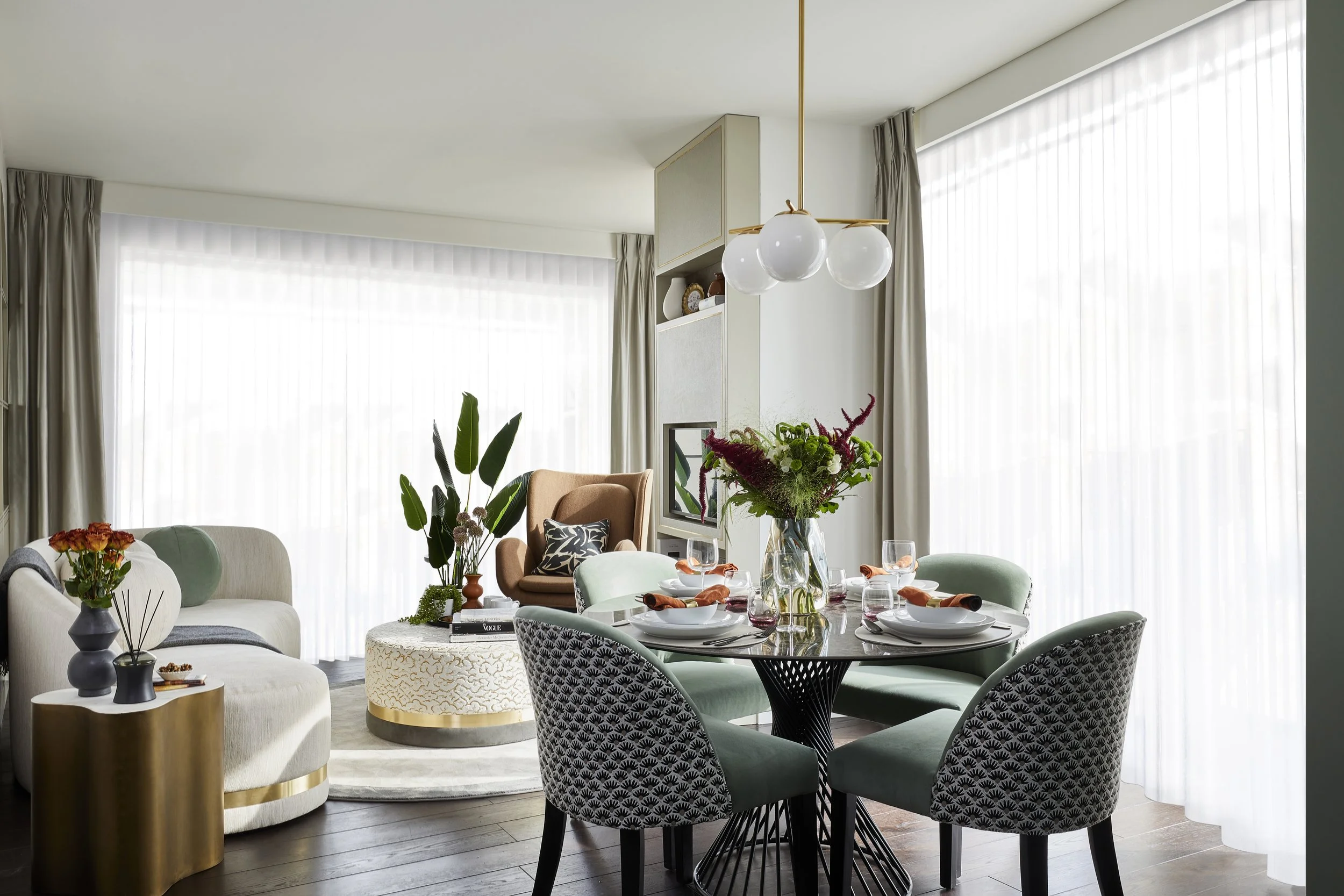 A modern dining area with a round table set for a meal, surrounded by patterned chairs, with decorative flowers in the center, large windows with sheer curtains, and contemporary lighting fixtures.