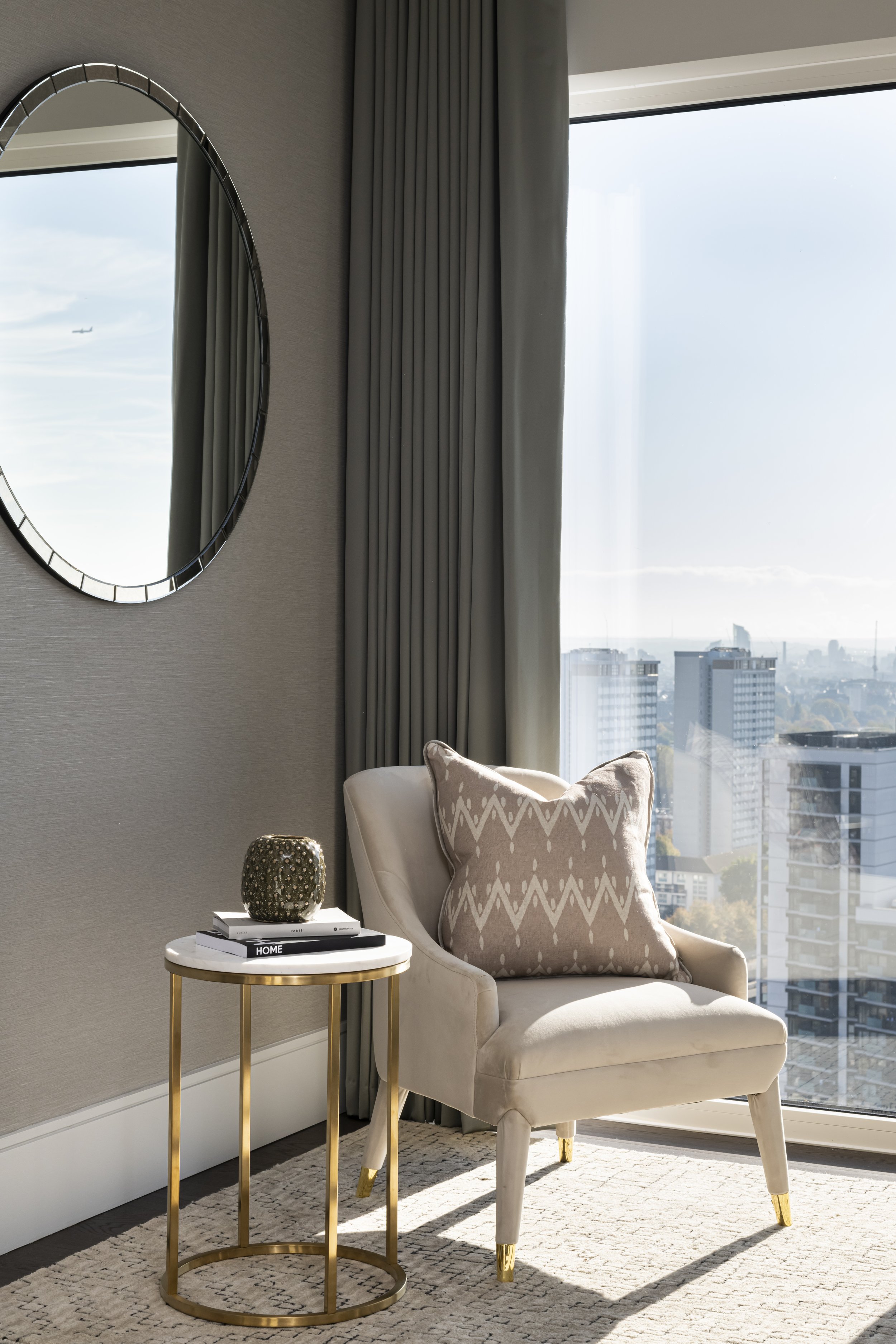 Interior scene with a cream-colored armchair, a side table with decorative objects, a mirror on the wall, and large windows with city view and curtains.
