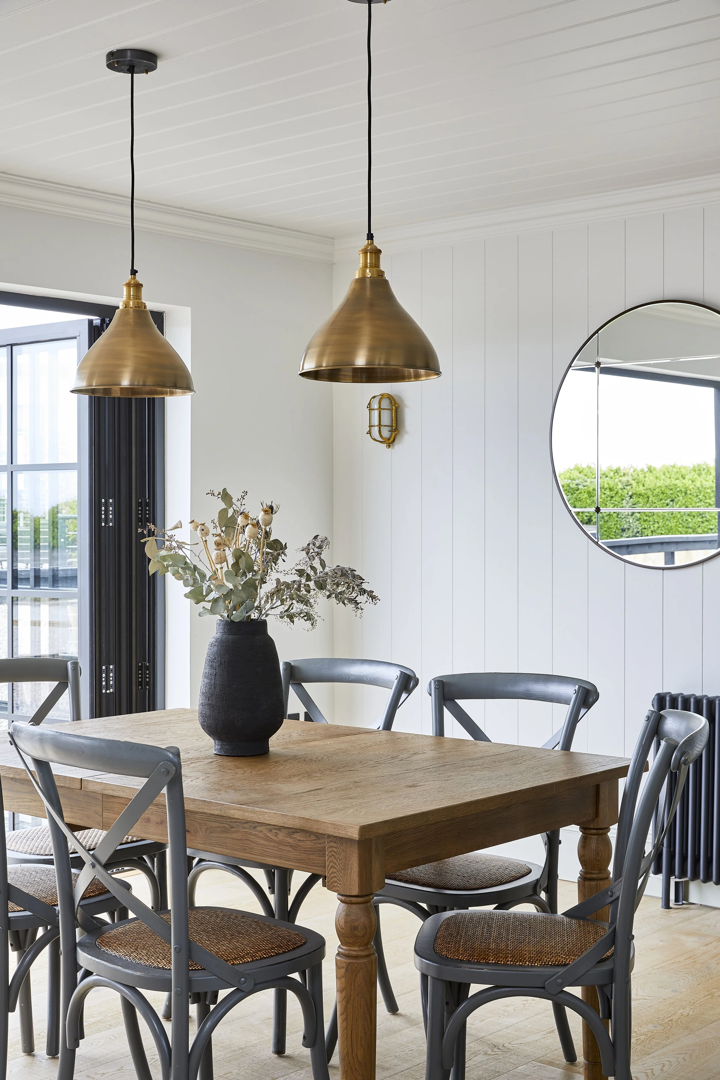 A dining room with a wooden table surrounded by chairs, a black vase with dried flowers on the table, two brass pendant lights hanging from the ceiling, a circular mirror on the wall, and a window with black framing showing a green landscape outside.