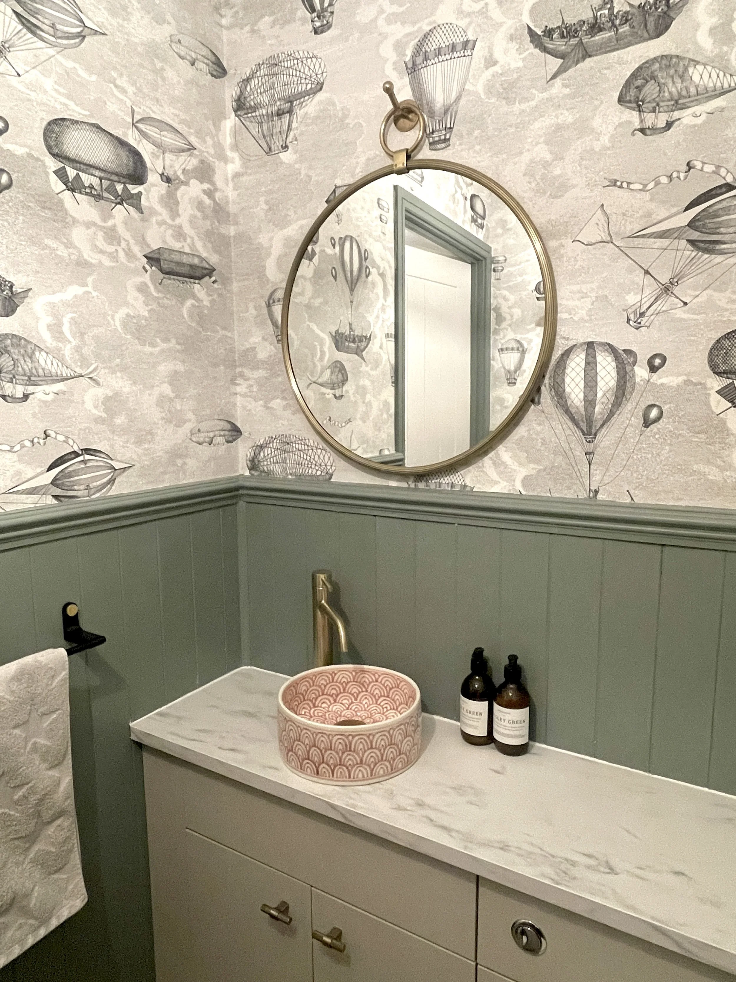 A bathroom vanity with a marble countertop, green lower cabinet, and a round gold mirror. The wall behind features wallpaper with airship illustrations. There are two bottles and a pink patterned basin on the counter, and a towel on a hook next to th