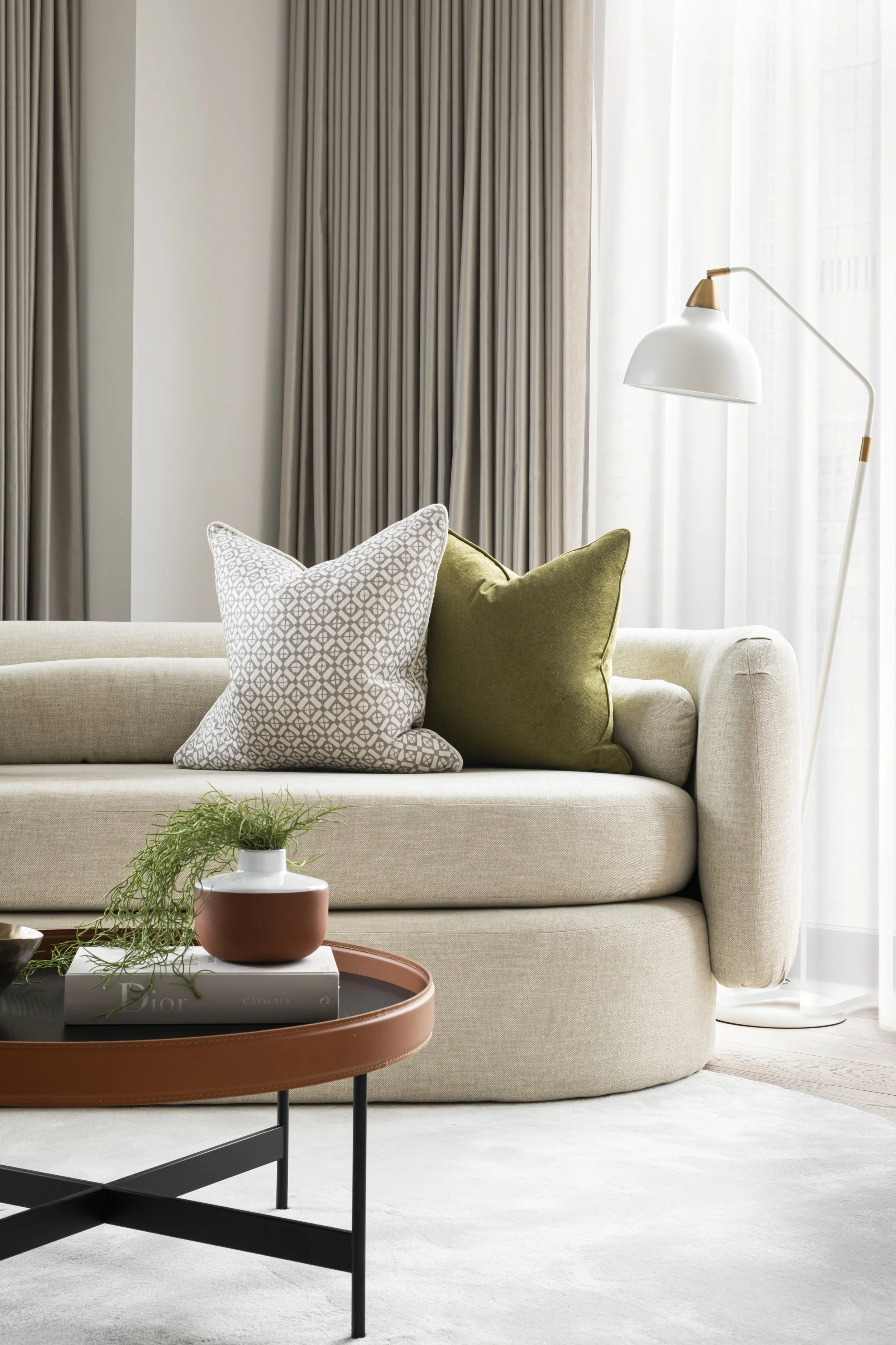 Living room with beige sofa, patterned and solid pillows, potted plant on coffee table, with tan tray and books, in front of curtains and a floor lamp near window.