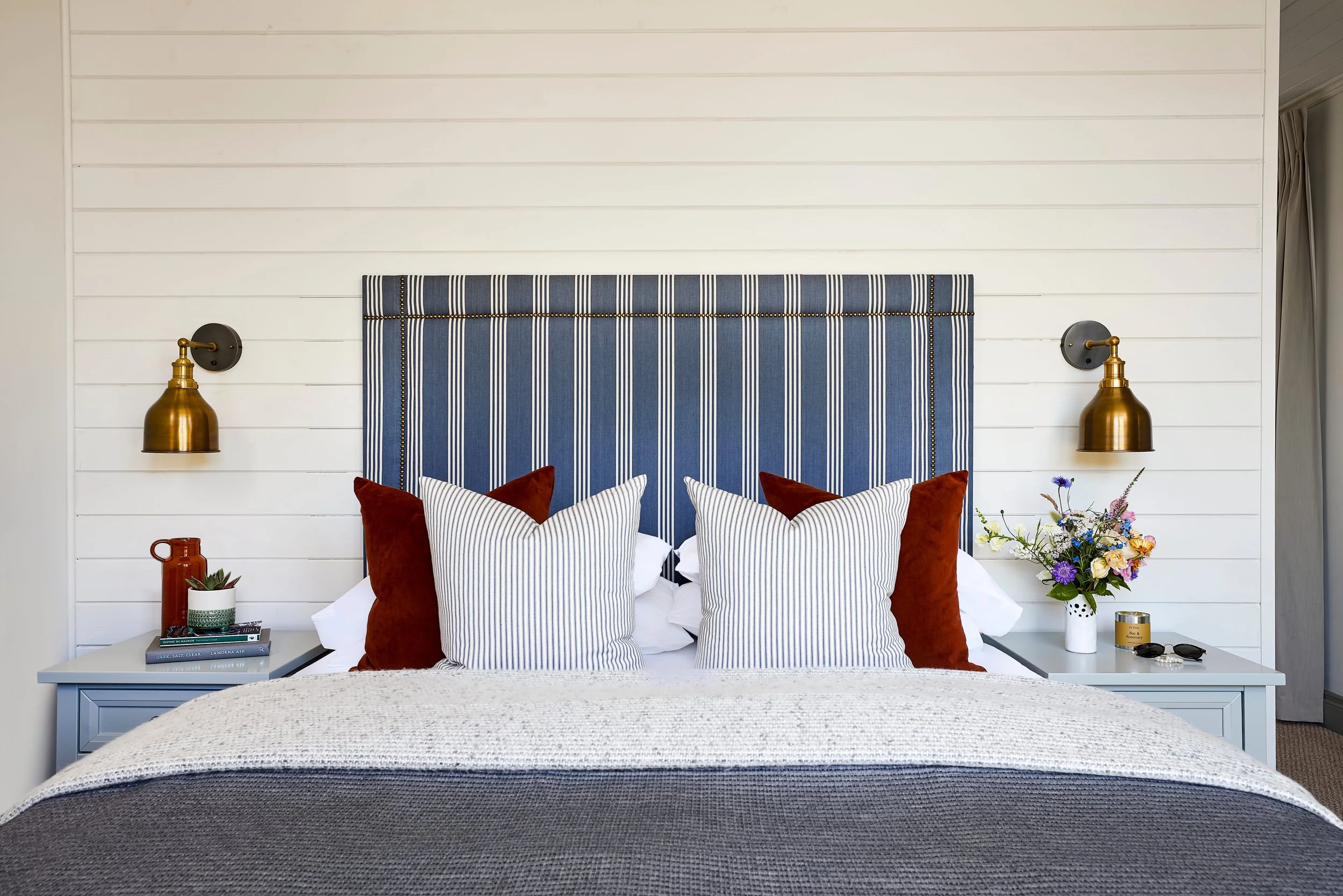 A neatly made bed with a navy blue and white striped headboard, decorated with assorted pillows including white, navy striped, and rust-colored pillows. The side tables hold a flowering plant, books, a mug, and decorative items. Wall-mounted gold and