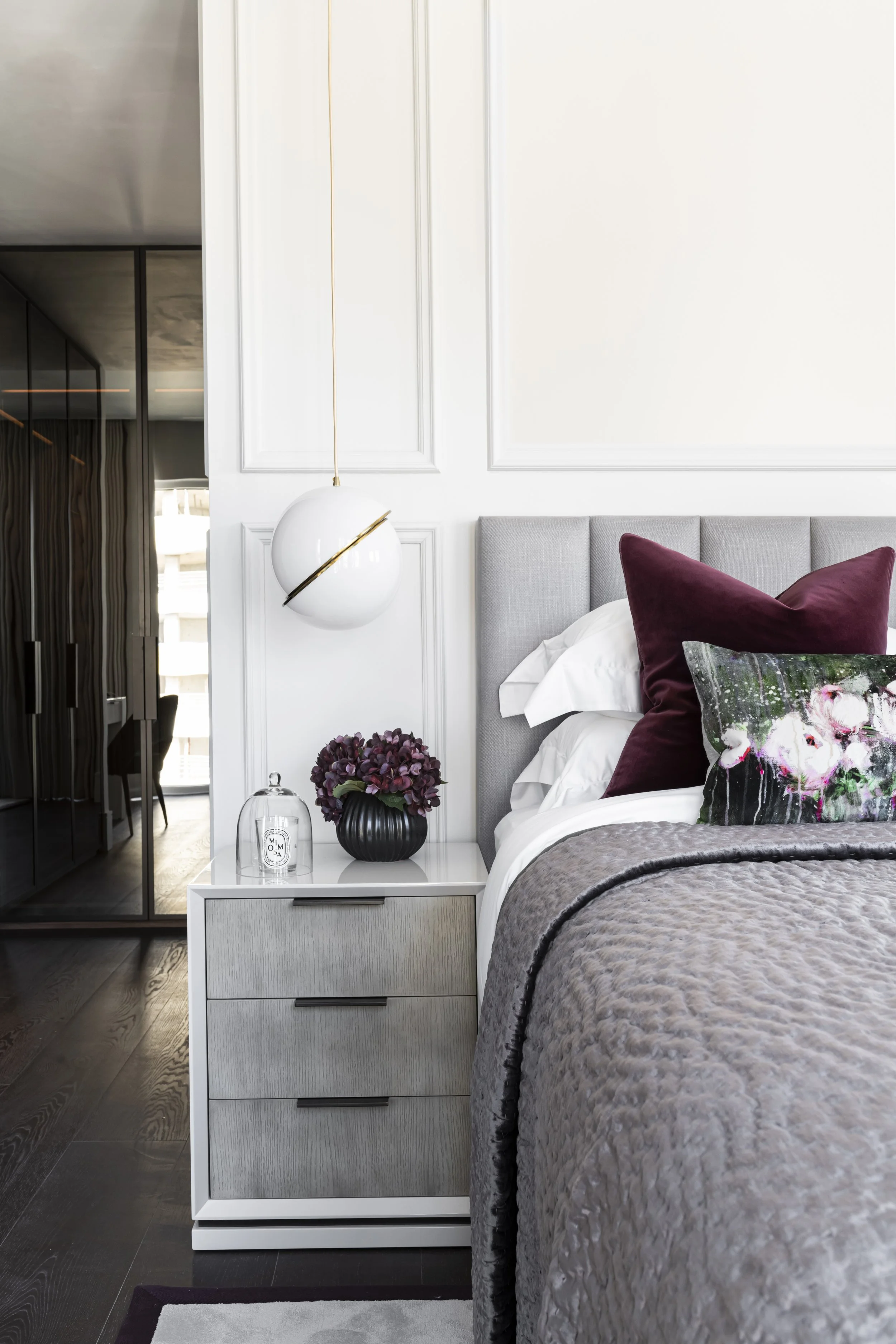 Modern bedroom with a bed and pillows, a white bedside with flowers, a hanging white and gold pendant light, and a wardrobe with sliding mirror doors.