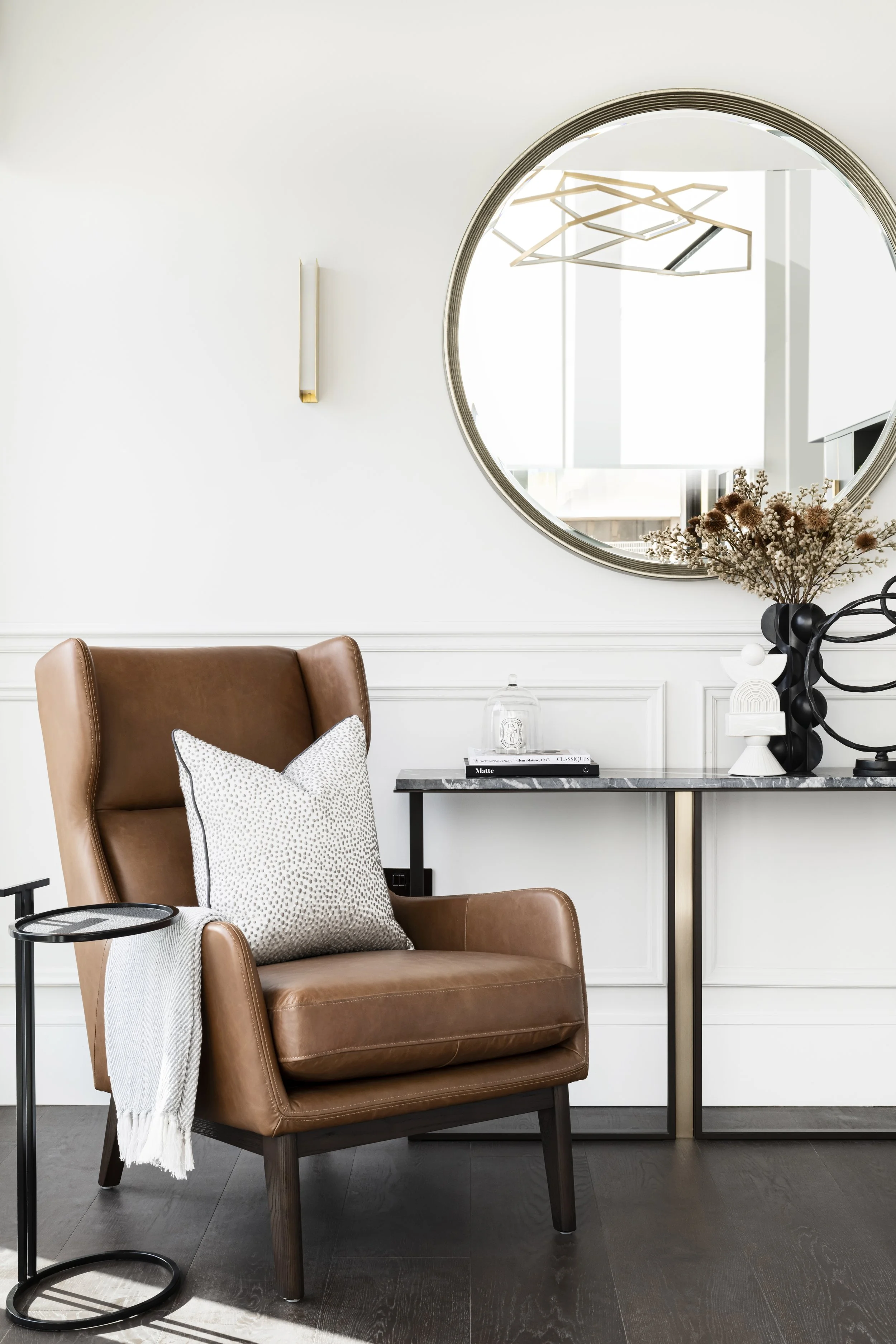 Living room interior with a brown leather armchair, a throw pillow, a side table with a glass dome, a white wall with a vertical light fixture, a round mirror, a black decorative sculpture, a vase with dried flowers, and a modern geometric chandelier