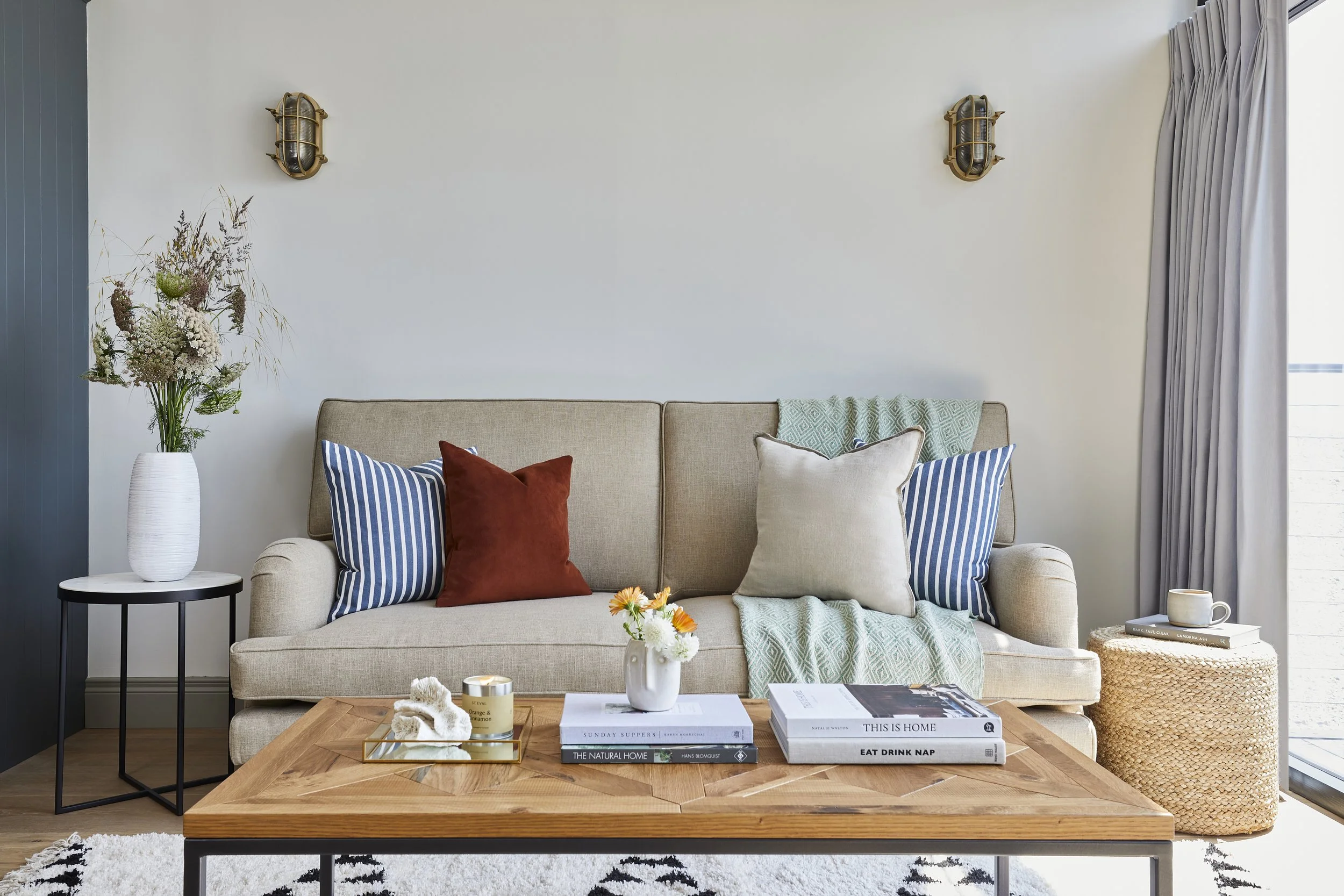 Living room with beige sofa, throw pillows, and coffee table decorated with books and a vase of flowers, near a large window with curtains and wall sconces.