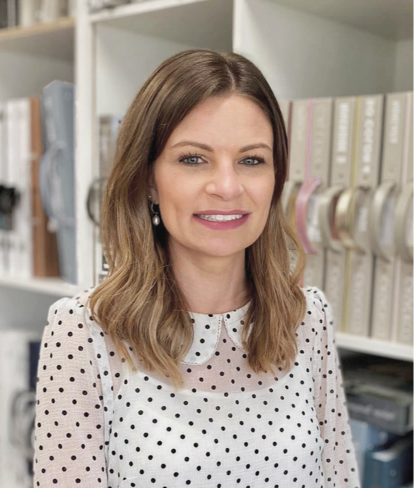 A woman with shoulder-length brown hair, wearing a white blouse with black polka dots and pearl earrings, smiling in front of a bookshelf filled with binders and books.