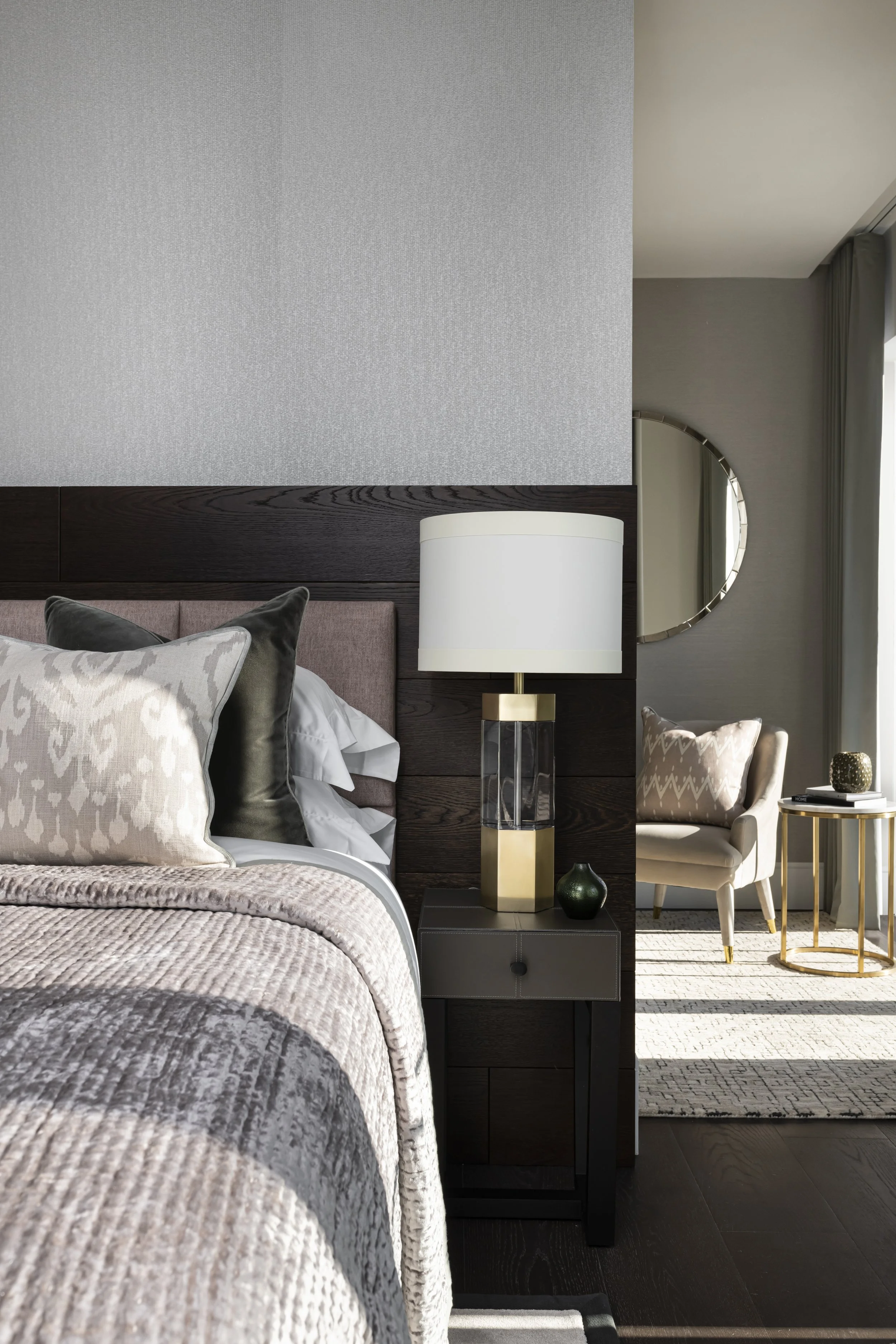 Close-up of a hotel bedroom with a bed, pillows, a bedside table with a lamp, and a sitting area with a chair and side table, and round mirror on the wall. Modern decor with neutral colors.