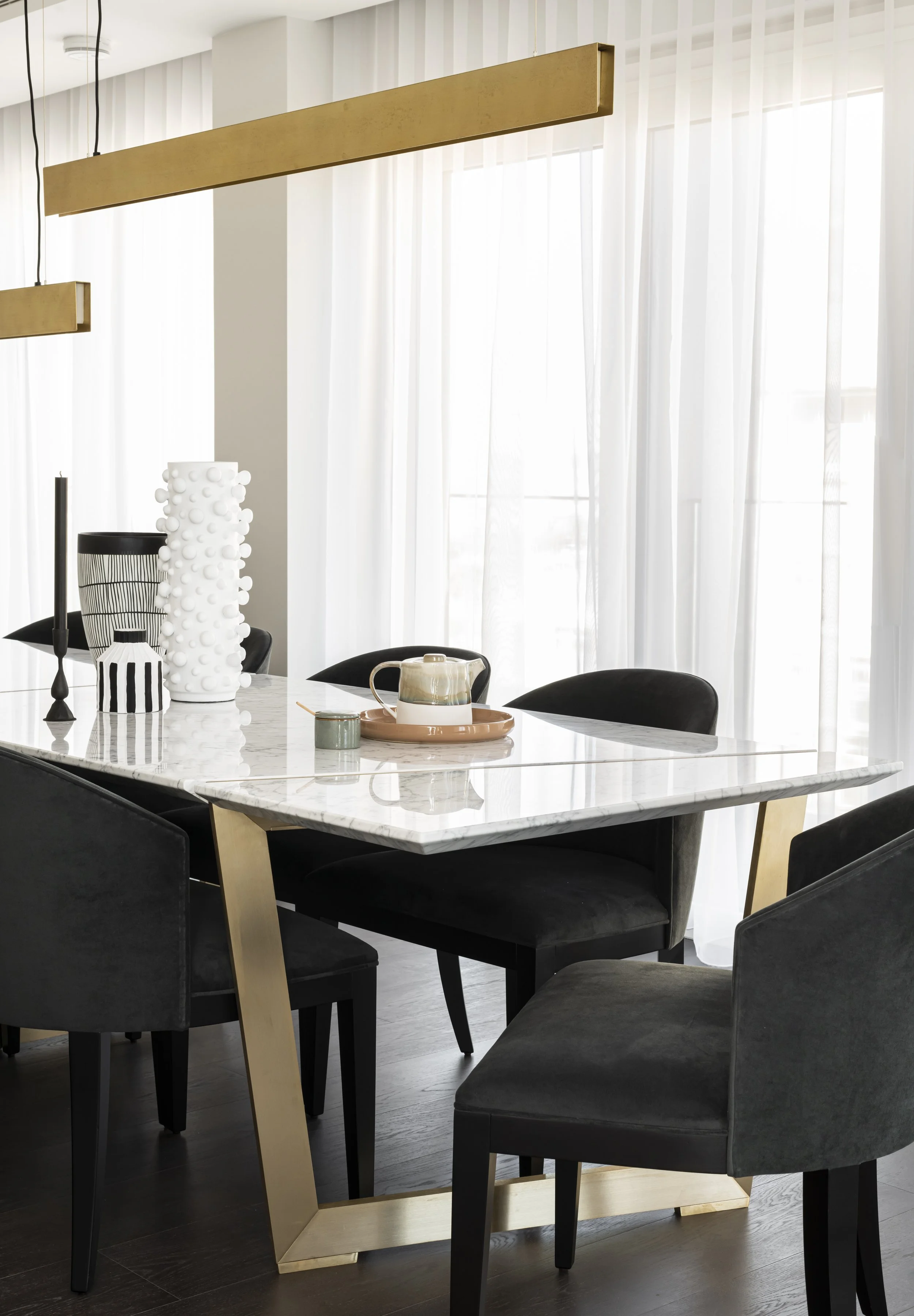 Modern dining room with a white marble table, black chairs, and decorative vases on the table, with large windows and sheer curtains in the background.