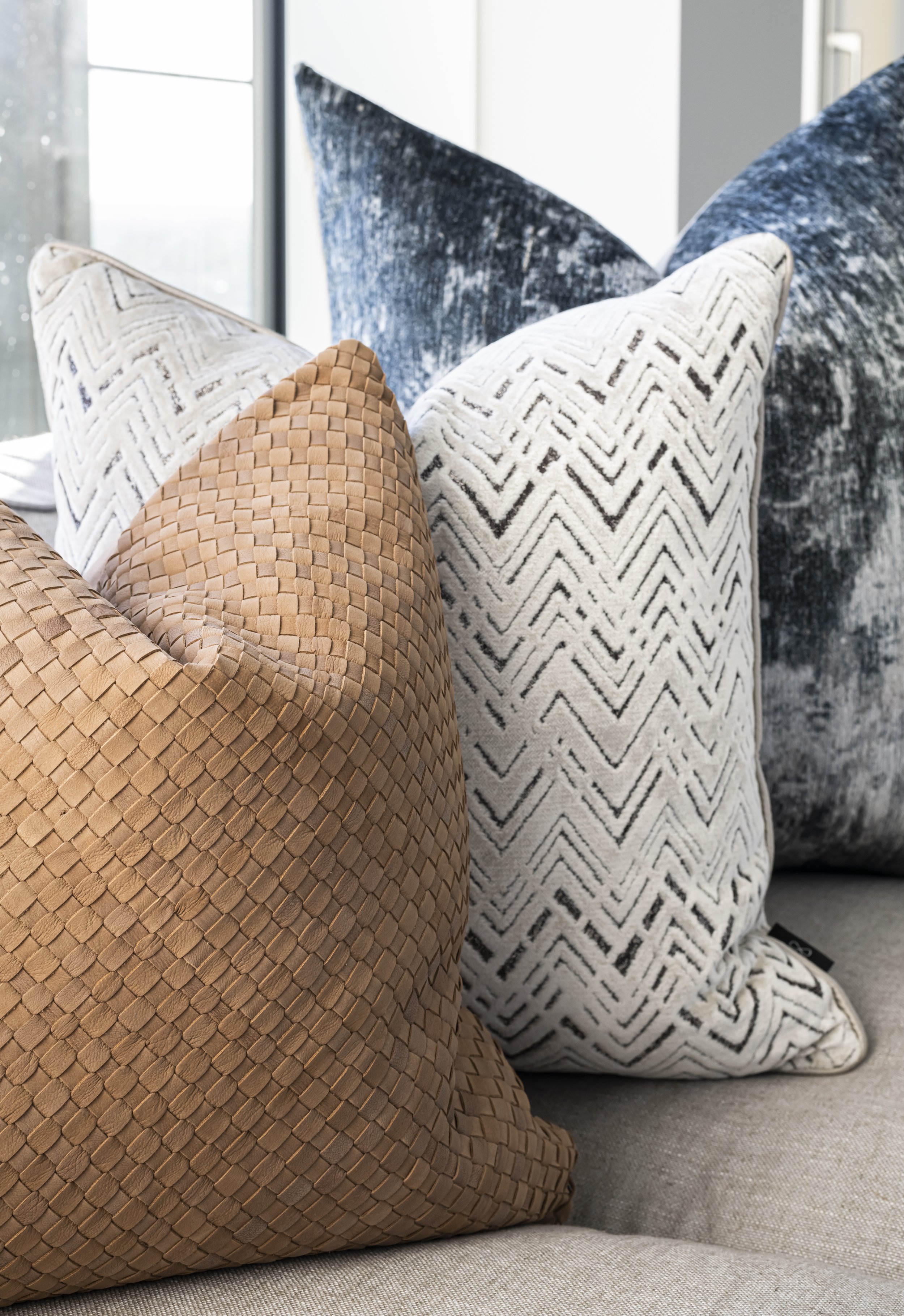 A collection of decorative cushions with various textures and patterns arranged on a neutral-colored sofa near a window.