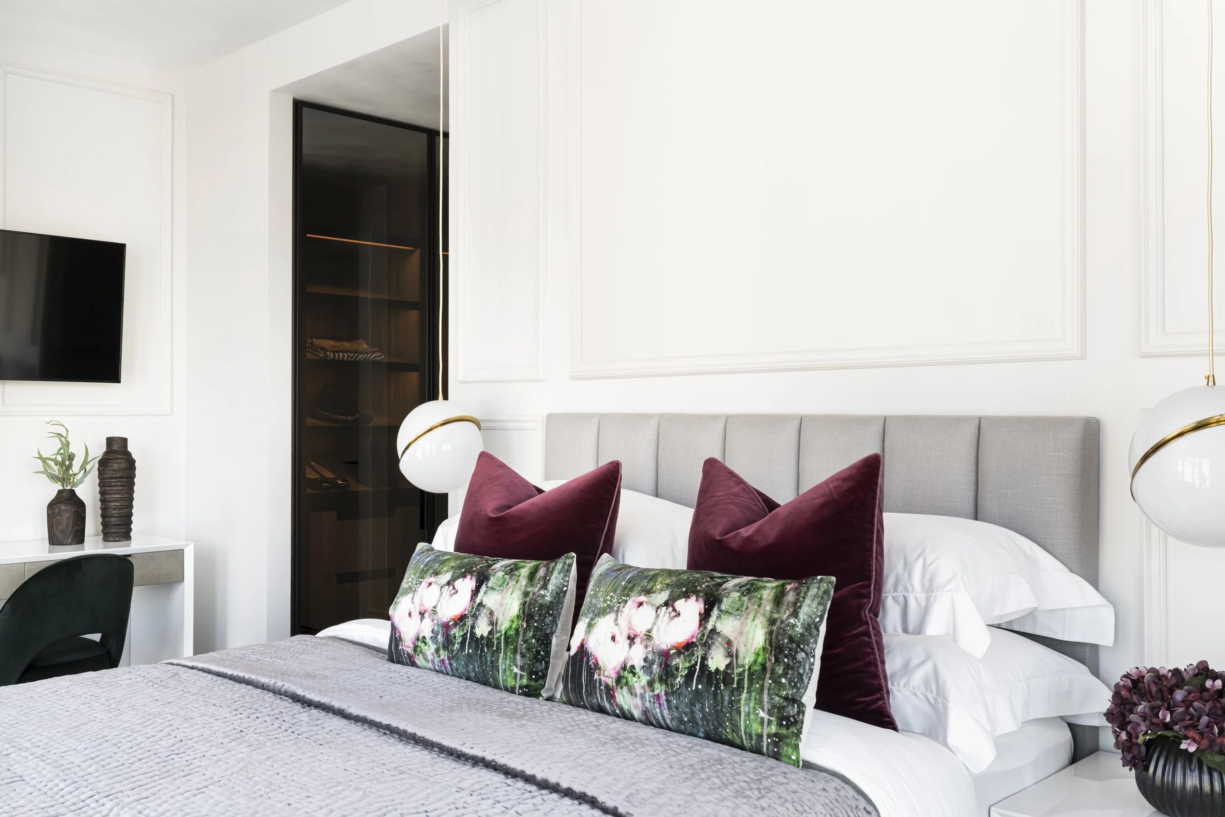 Modern bedroom with a bed decorated with several colorful and velvet cushions, including floral patterned pillows, against white walls with decorative moulding, a wall-mounted flat-screen TV, and a black wardrobe door with glass panels.