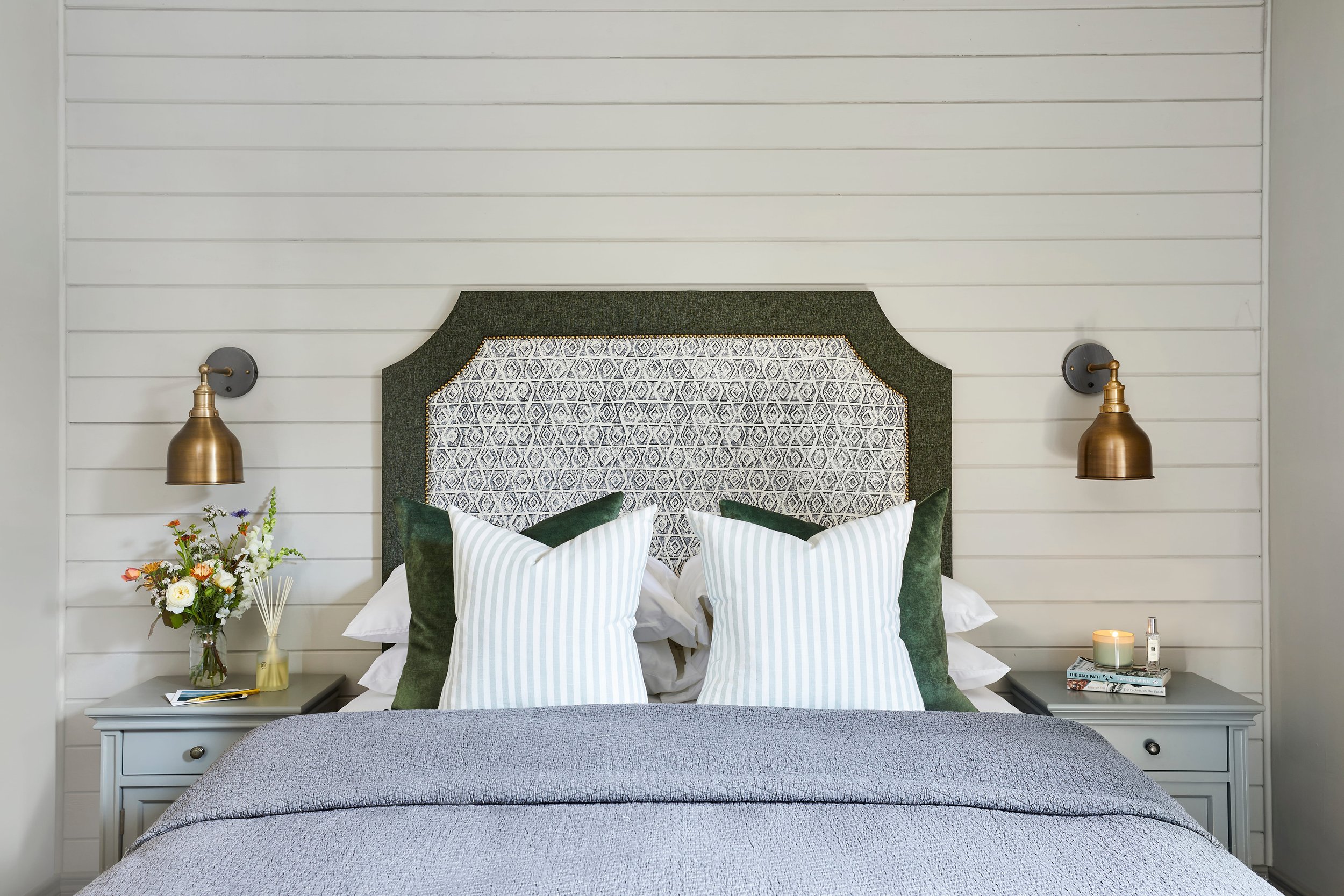 A neatly made bed with a patterned headboard and striped pillows in a bright bedroom. Flower arrangements and a few books are placed on nightstands on either side of the bed, with wall-mounted lamps above each.