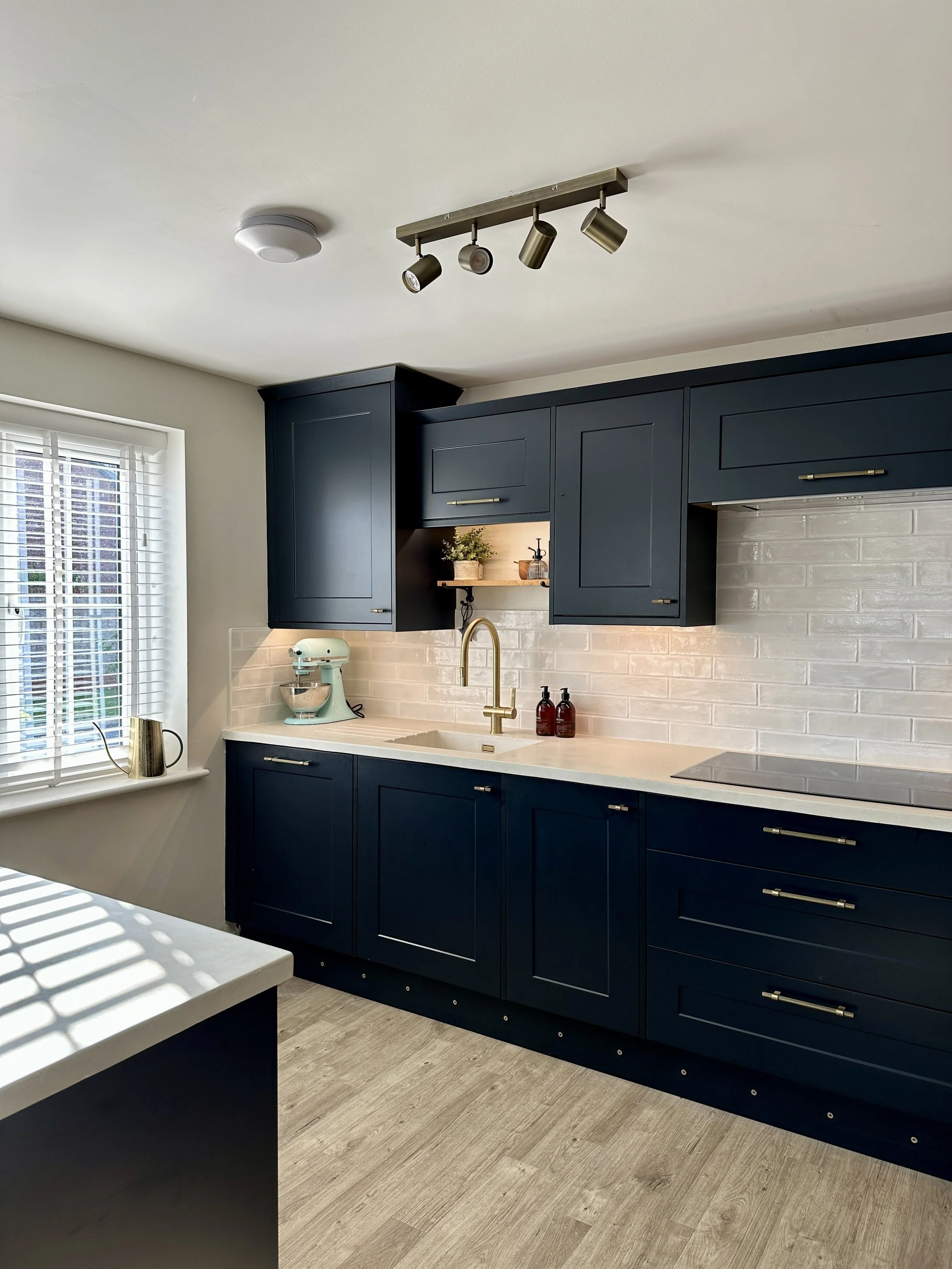 A modern kitchen with navy blue cabinets, a white countertop, a white brick backsplash, a gold faucet, and a window with blinds. There are pendant lights and a small open shelf with a plant and decorative items.