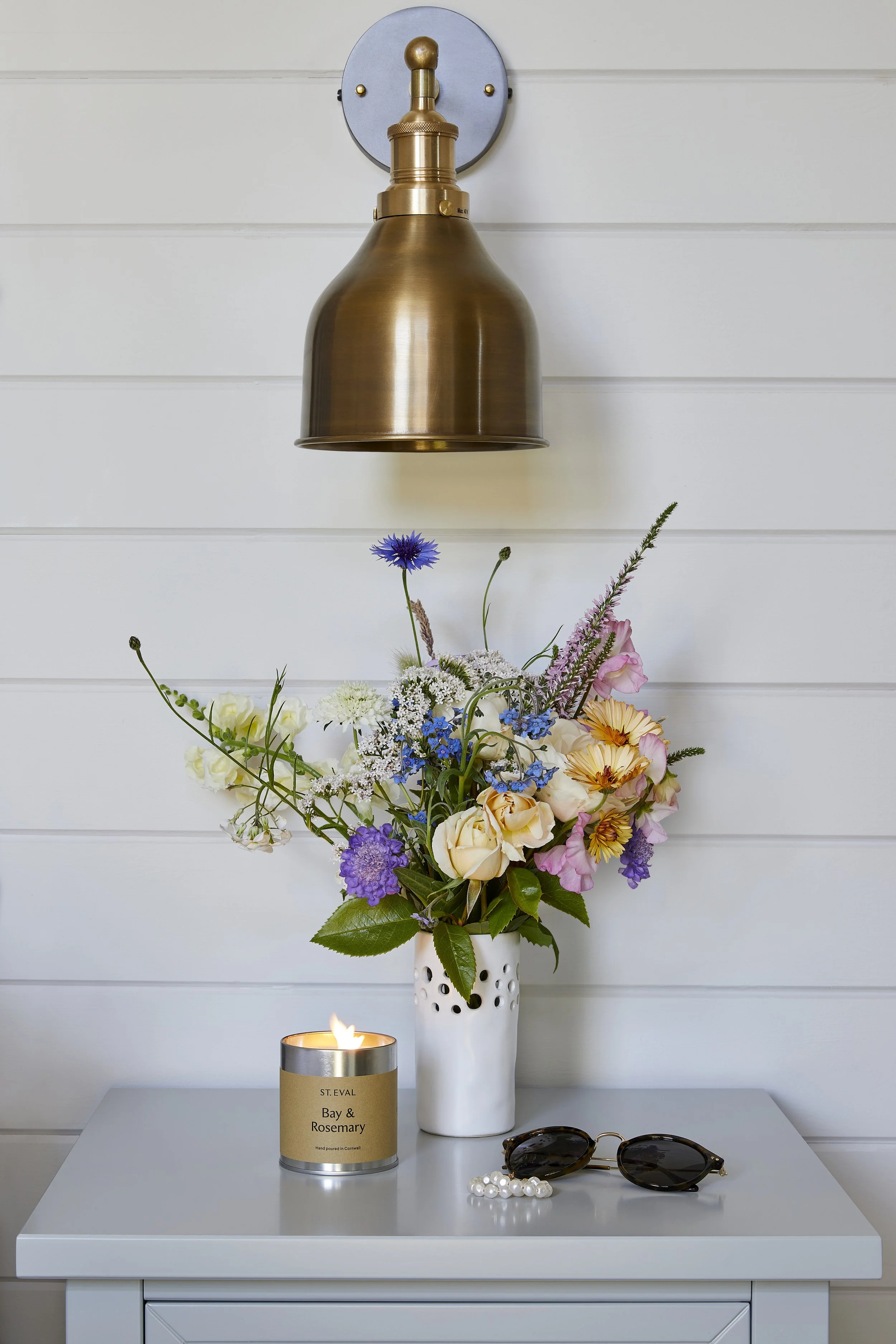 A  side table with a tall white vase holding a colorful flower arrangement, a lit candle in a gold and silver container labeled "Bay & Rosemary" placed in front, and a pair of sunglasses and pearl bracelet on the table. A brass wall-mounted light fix