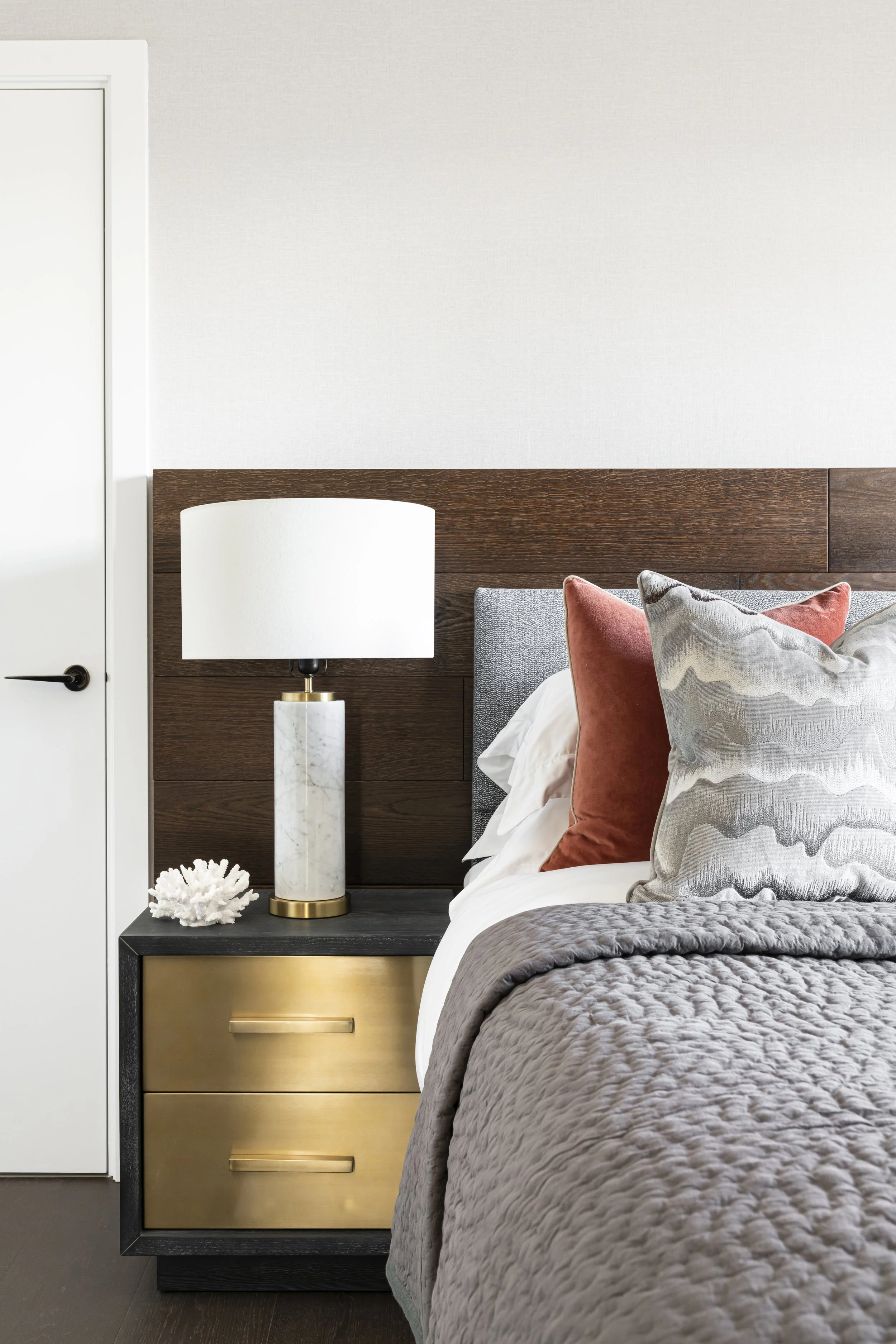 Close-up of a bedroom corner showing a bedside with a marble-based lamp, decorative coral, and plush cushions on the bed with textured grey and rust-coloured fabric. The background features a dark wooden headboard and a white wall.
