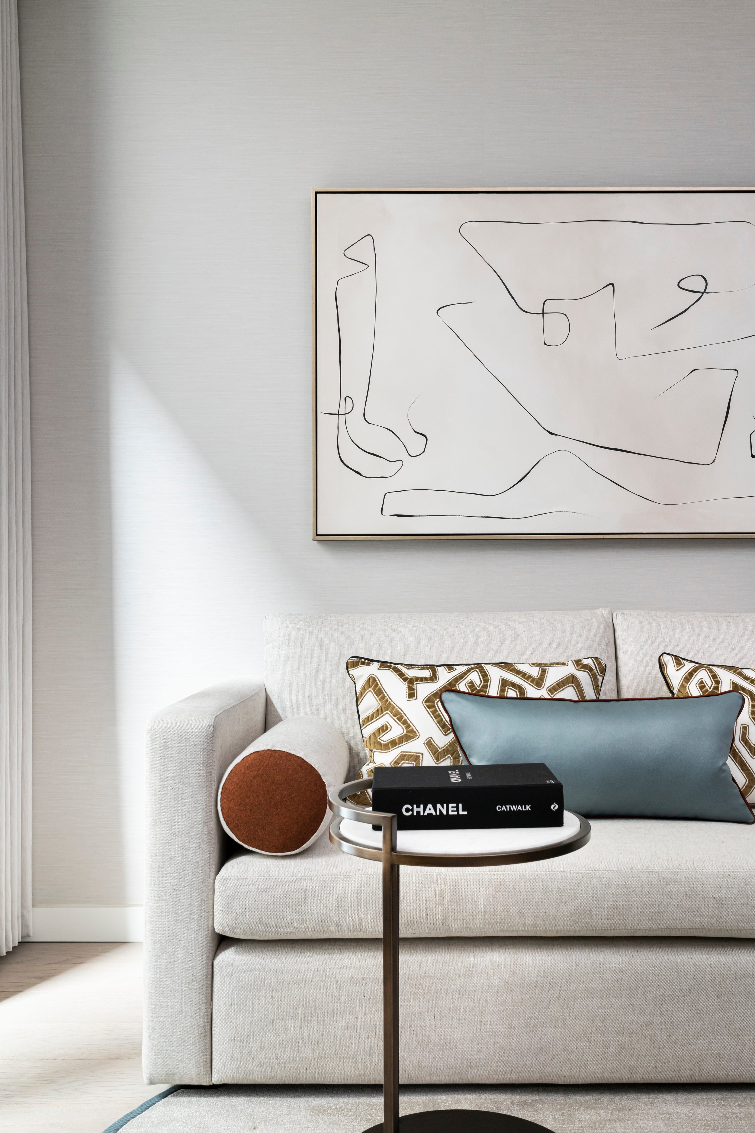 A modern living room corner with a neutral sofa, decorative cushions, a side table with a Chanel book, and a large abstract line art painting on the wall.