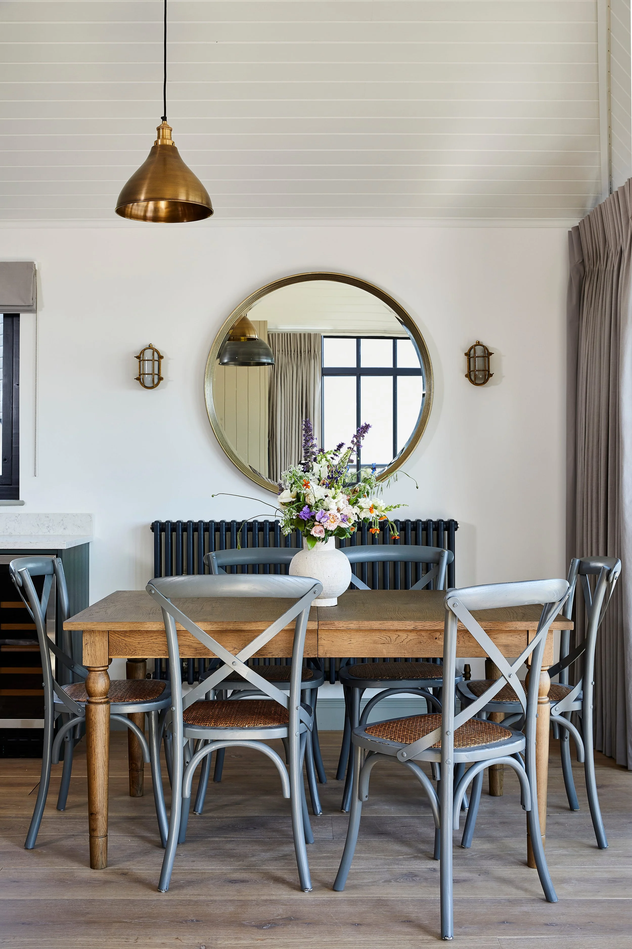 A dining room with a wooden table, gray chairs, a white vase with flowers, a round mirror on the wall, and a window with curtains. A brass pendant light hangs from the ceiling.