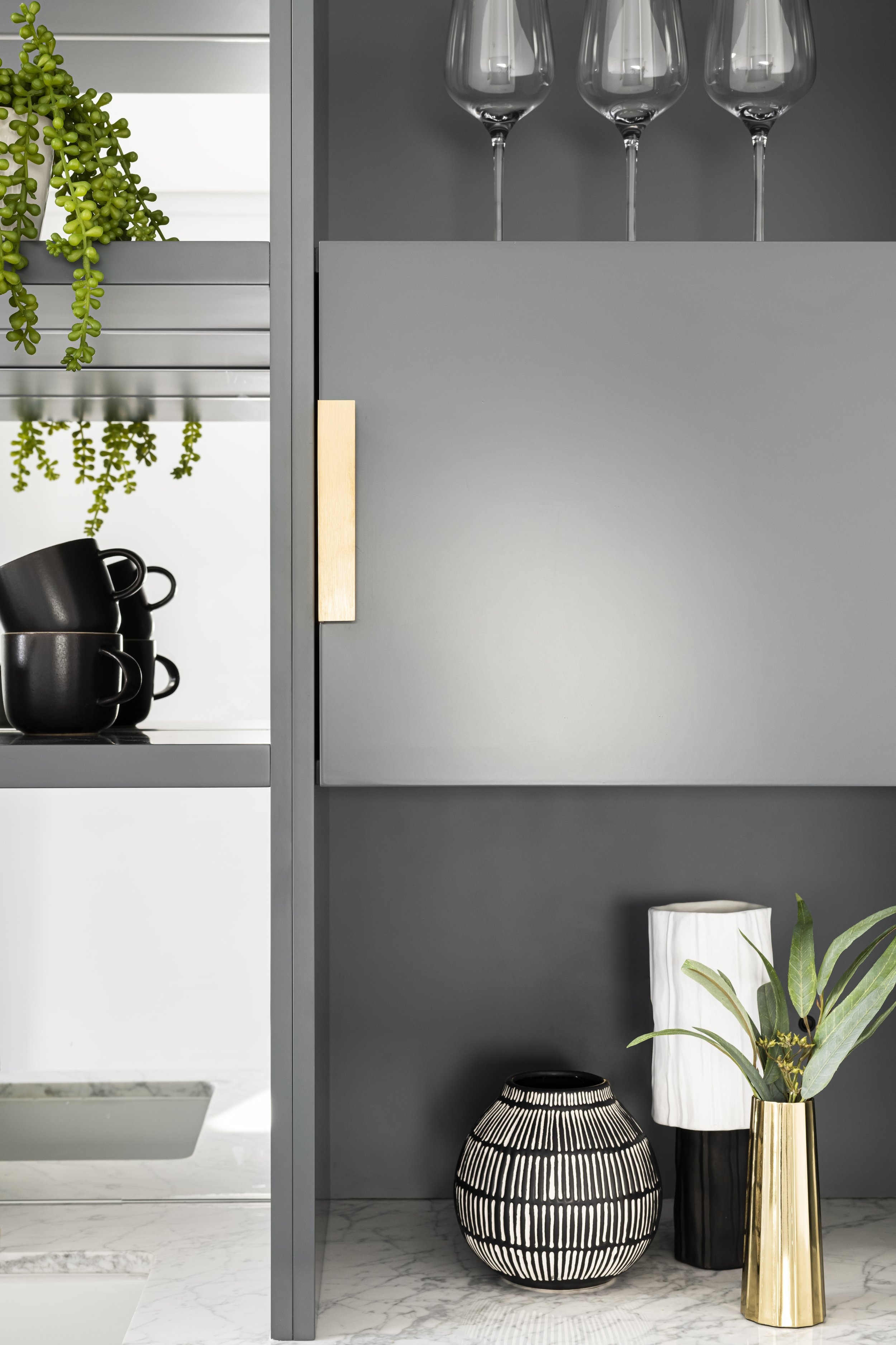 A black and white striped decorative vase, a tall white vase with green plants, a black and gold vase, and a black globe-shaped lamp with white lines, all placed on a marble floor next to a dark gray wall.